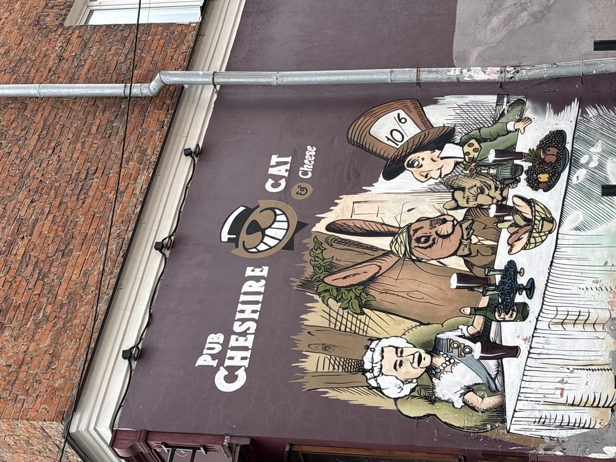 The image shows a mural for the "Pub Cheshire Cat & Cheese". The mural depicts characters resembling those from Alice in Wonderland sitting at a table, including the Mad Hatter, the March Hare, and a person resembling the Queen of Hearts. The mural is painted on the exterior wall of a building, adjacent to a brick building. The building has a downspout running horizontally across the top of the mural. Based on previous information, this mural is located in Perm, Russia.