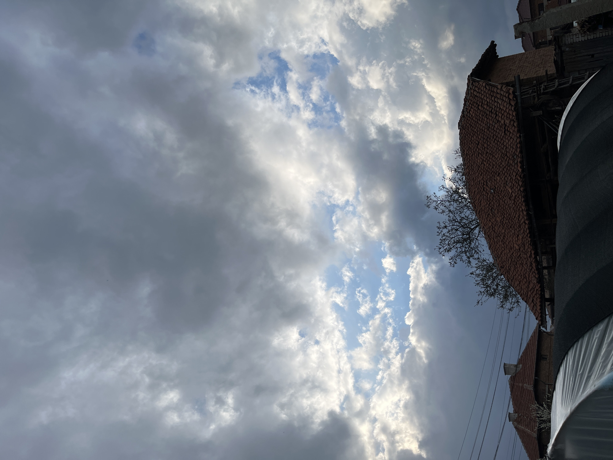 The image shows a cloudy sky above a structure, possibly a building or house, in Karavelovo, Bulgaria. The sky is filled with a mix of dark gray and white clouds, with patches of blue sky visible in between. The structure has a red tiled roof, a bare tree is visible, and a black cylindrical object and electrical wiring are in the scene.