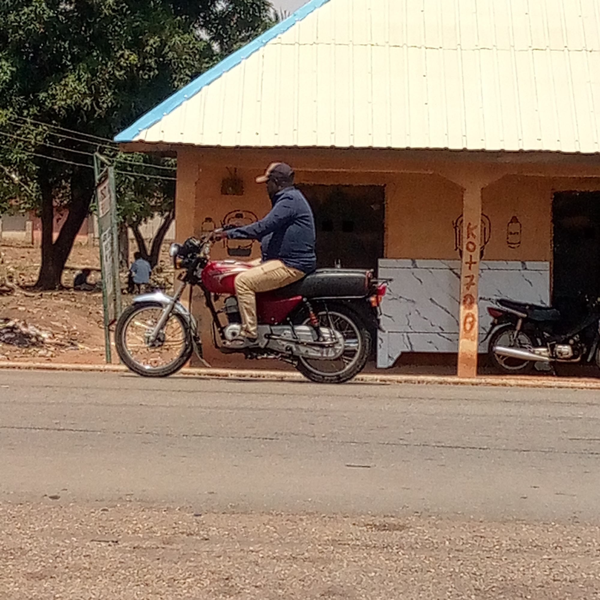 A man is sitting on a maroon motorcycle parked on a paved road in front of a building with a tan exterior and a light-colored roof with a blue trim. The man is wearing a dark blue long-sleeved shirt, tan pants, and a baseball cap. Behind the motorcycle, another motorcycle is parked near a building post with some writing in an unidentifiable language. In the background, there is a tree and two individuals seated on the ground. The location is in the city of Unknown, Unknown.