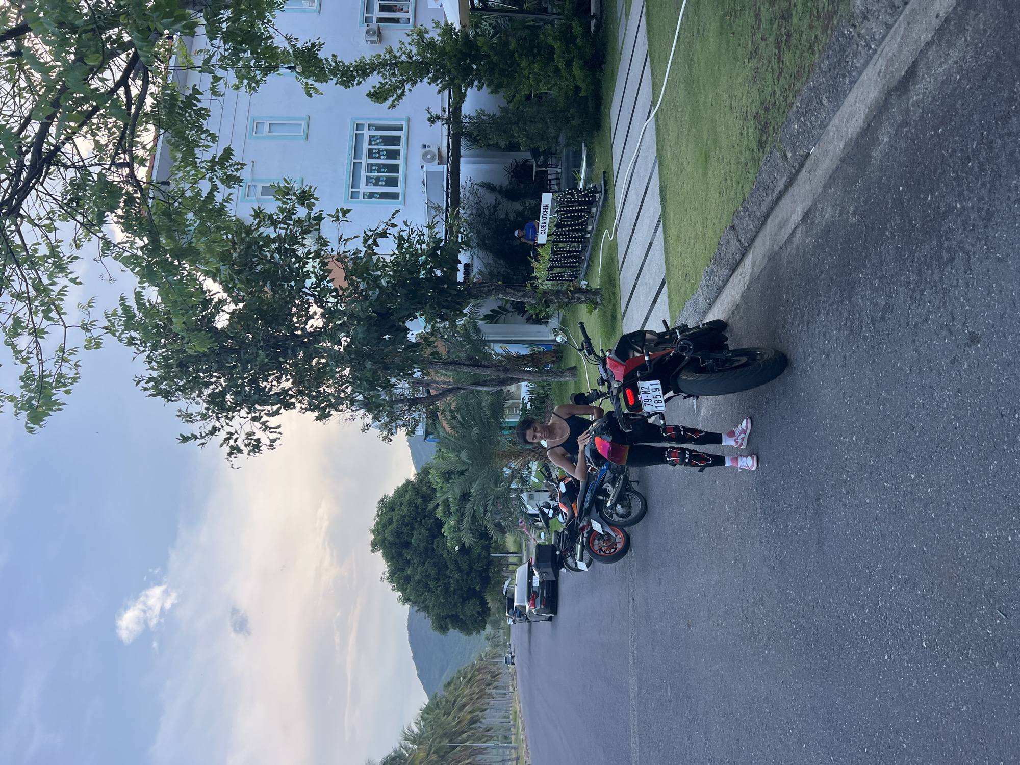 A woman is standing on a roadside near several parked motorcycles. She is wearing a black top, dark pants, and pink and white sneakers. One of the motorcycles behind her has a license plate reading "79-M2 185.91". The location is near a building with light blue trim and what appears to be a cafe or restaurant sign that reads "ANANDA CAFE & KITCHEN". The environment includes a paved road, a grassy area, and trees, likely located in Nam Nha Trang Ward, Vietnam.