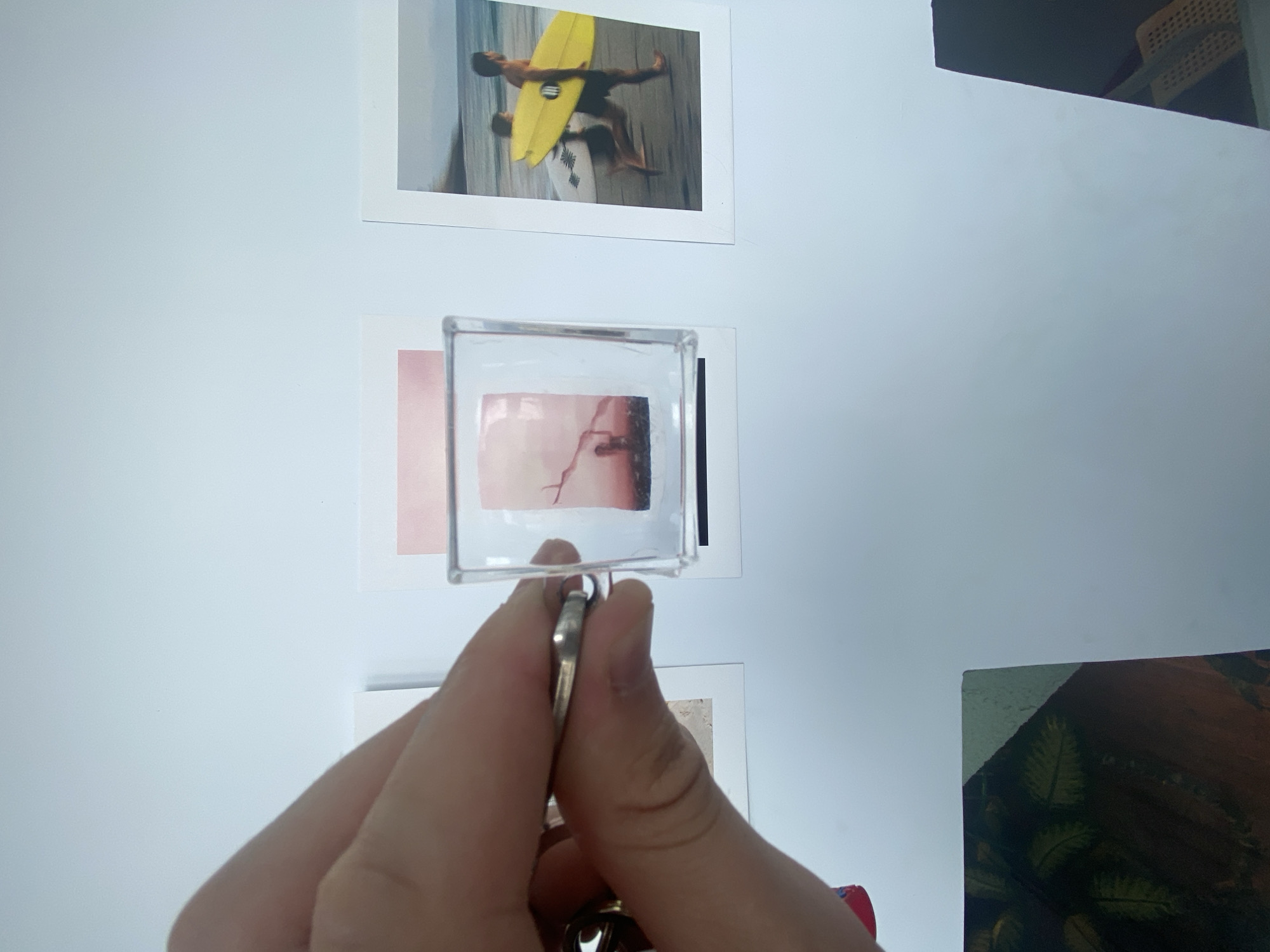 A hand is holding a clear, square plastic frame with a small pink and red toned image inside. The image depicts a silhouette of a person standing under a bare tree against a sunset or sunrise sky. The frame is suspended from a keyring. Behind the frame, three photos are affixed to a white surface. The leftmost photo is partially visible. The middle photo is a polaroid with a pink wash at the top and a silhouetted person under a tree. The rightmost photo is a polaroid of two people running on a beach, one carrying a yellow surfboard. The overall scene suggests a collection of personal mementos displayed on a wall. There is no discernible information within the image that would indicate a specific location in Khabarovsk, Russia.