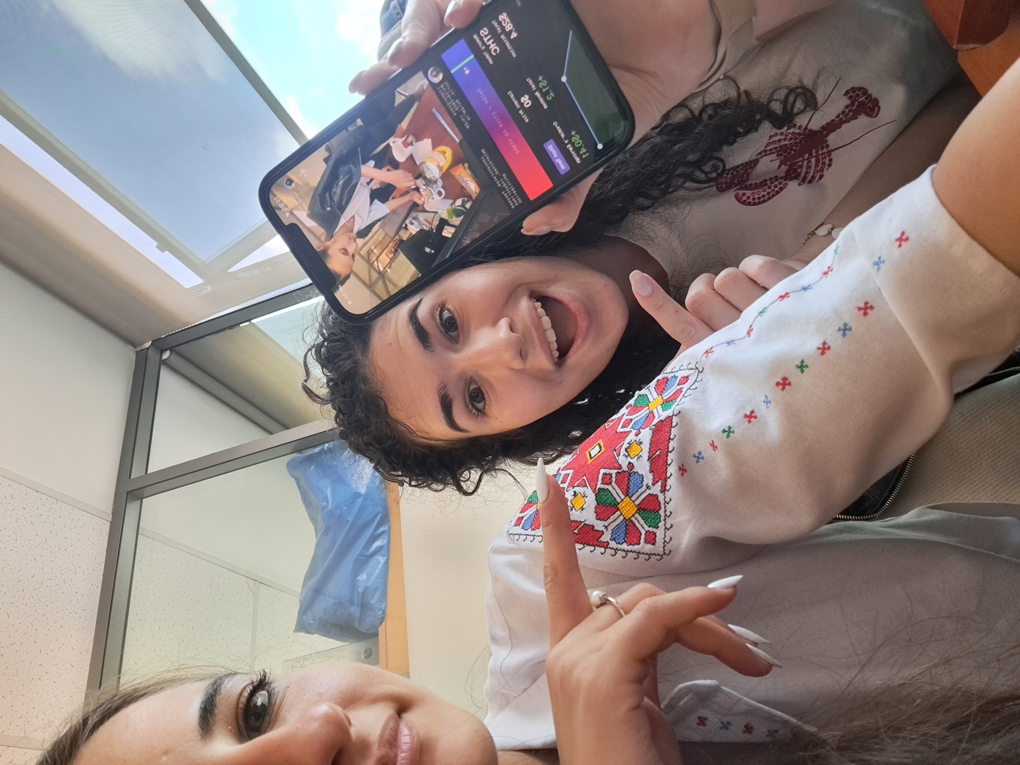 A vibrant, high-angle indoor selfie captures two young women in a casual setting. The woman in the foreground has dark, curly hair and a wide, joyful smile, with her mouth open. She is pointing with her right hand, showcasing long, white, pointed nails and a ring on her ring finger. She wears a white top featuring colorful, geometric embroidery on the sleeve, reminiscent of a traditional vyshyvanka, and a distinct red lobster graphic on the main body of the shirt.
She holds a black smartphone upside down, displaying a screen with various text and numbers, including "2584", "+201", "+21'5", "20", "met horie", "jores pereune", "CTLLBNE BLICS", "Buriciba1100", "GREBLUTUNG", "COUCADEL", "оспаев", and "esar". The phone's screen also shows an image of a person at a table with mugs and other items.
Part of a second person's face, including an eye, is visible in the bottom left corner. The background features bright daylight streaming through an overhead window or skylight, suggesting it is daytime with a clear to partly cloudy sky. Below the window, glass partitions and a dropped ceiling indicate an indoor environment, possibly an office or modern communal space. A blue object is visible behind the second person. The overall mood is cheerful and candid.