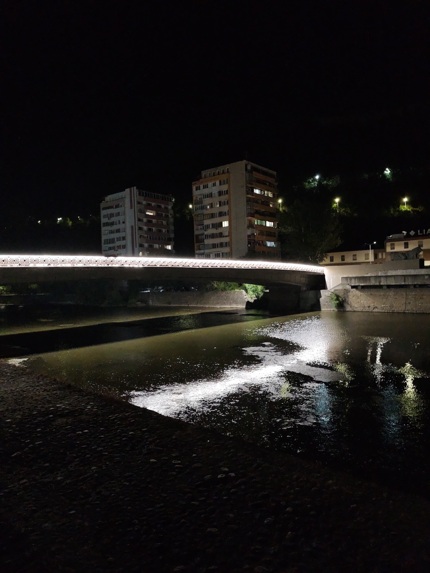 The image depicts a night scene of the Old Bridge in Lovech, Bulgaria, spanning the Osam River. The bridge is illuminated with a string of lights that creates a patterned glow along its railing. On the left side of the bridge, a tall, light-colored residential building with visible windows is visible. To the right of this building and further in the background, another similar residential building stands, also lit from within. Beyond the buildings, the dark silhouettes of trees and a hillside are present, with several streetlights casting pools of light. The river below reflects the lights from the bridge and the buildings, creating shimmering patterns on its surface. The foreground shows a dark, textured embankment, likely made of cobblestones or similar material.