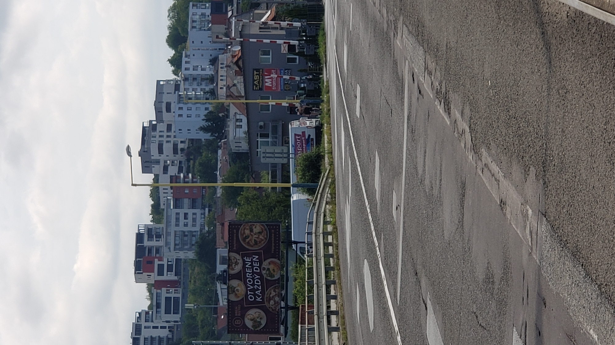 The image captures a street-level view of an urban environment, likely Prešov, Slovakia, with a focus on modern architecture and infrastructure. In the background, a cluster of contemporary apartment buildings, characterized by their geometric designs and a mix of white, grey, and red facades, are situated on a hillside. A bright yellow street lamp stands prominently in the middle ground, its pole a stark contrast against the overcast sky.
To the left, a large billboard displays advertisements for "Brio Bistro" and features images of pizzas and other dishes, with the text "OTVORENÉ KAŽDÝ DEŇ" (Open Every Day) prominently featured. Adjacent to the billboard, a white van with "MV transport" branding is visible.
The foreground consists of a paved road with visible road markings, including pedestrian crossing lines, indicating a street intersection or a crossing area. The road surface shows signs of wear and tear, with several patches and cracks. A metal guardrail runs along the side of the road, separating it from a possibly elevated area or bridge. In the far right of the image, a railway crossing with striped barriers is partially visible, suggesting proximity to a train track. The overall atmosphere is that of a functional urban area on a cloudy day, with elements of residential housing, commercial advertising, and transportation infrastructure.