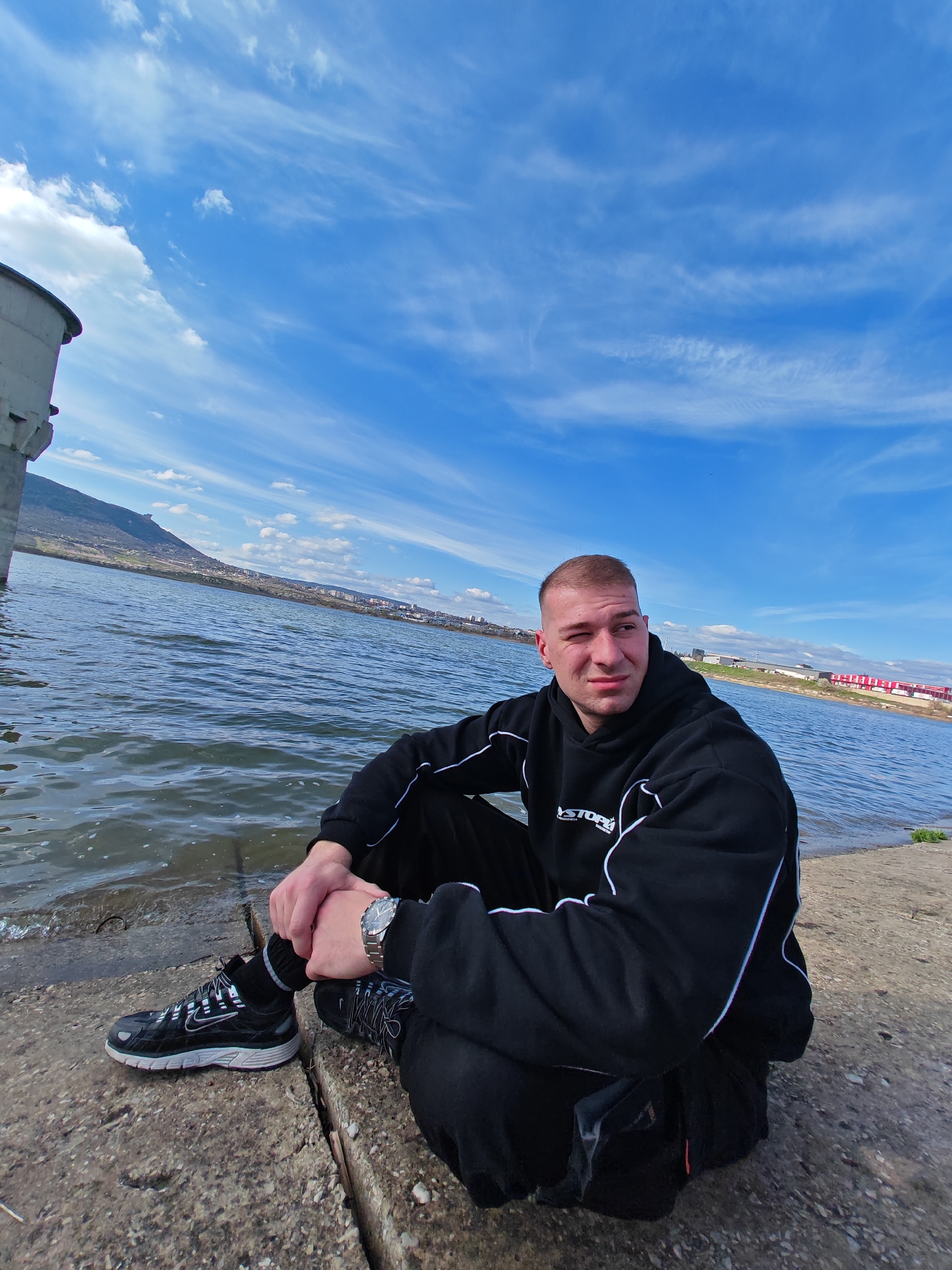 A young man with short hair is sitting on a concrete embankment next to a body of water, which may be the reservoir called "Yazovir Koshukar," with a view of the city of Shumen, Bulgaria, in the background. He is wearing a black hoodie with white stripes down the sleeves, black pants, and black athletic shoes. He also wears a wristwatch on his left wrist. The sky is blue with scattered clouds. In the distance is a mountain range and a row of red-roofed buildings. Part of a large cylindrical structure is visible on the left side of the frame. The water has small waves.