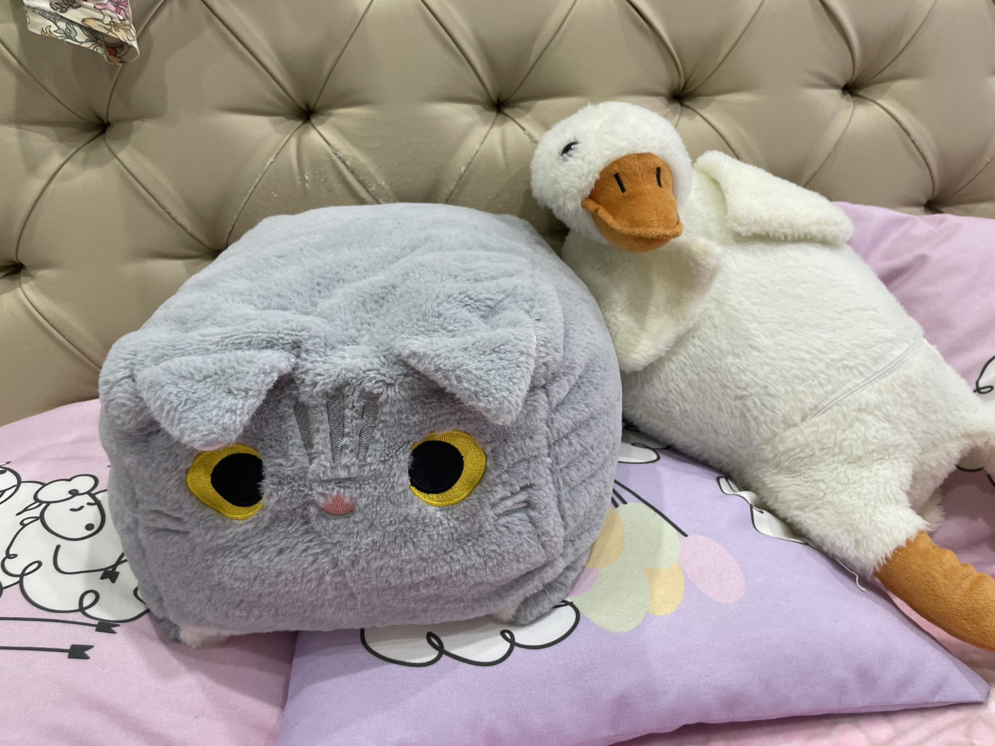 A gray, cat-shaped plush pillow and a white, duck-shaped plush toy rest on a pink and purple pillow adorned with drawings of sheep and clouds. The backdrop is a padded, beige headboard. The location of the scene is not discernible from the image.