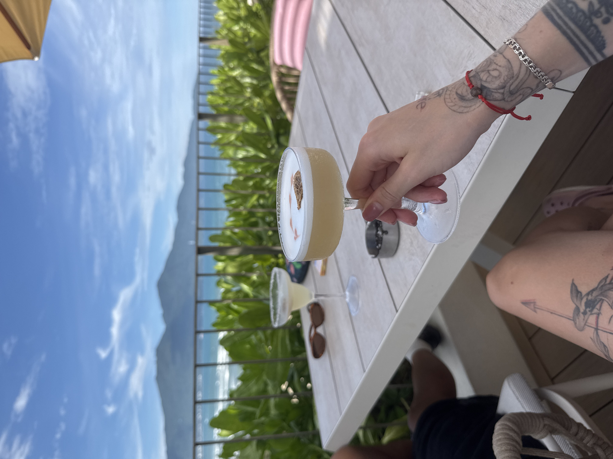 A person sits at a table on a balcony overlooking a beach in Đà Nẵng, Vietnam. The person is holding a cocktail with a frothy top and a garnish in one hand. On the table, there is another cocktail, sunglasses, and an ashtray. The person has tattoos on their arms and legs and is wearing a red bracelet. The balcony has a metal railing, and the view includes the ocean, a beach, and a mountain in the distance. The sky is partly cloudy.