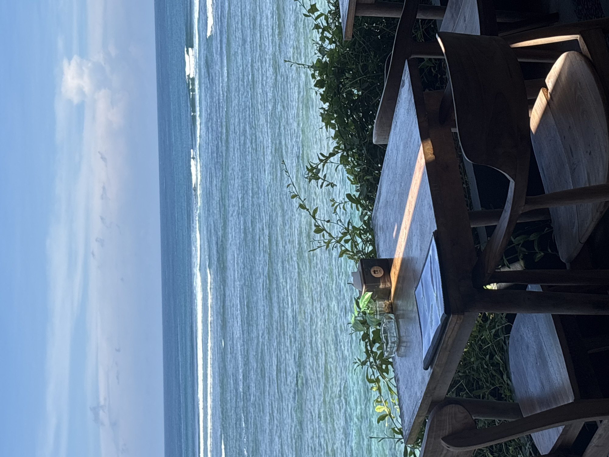 The image shows an outdoor dining area with a view of the ocean in Kuta Selatan, Indonesia. A wooden table and chairs are positioned next to lush green foliage, overlooking the water. The ocean is turquoise with white-capped waves. The sky is blue with a few wispy clouds. A menu and some small decorative items are on the table. There are no people visible in the image.