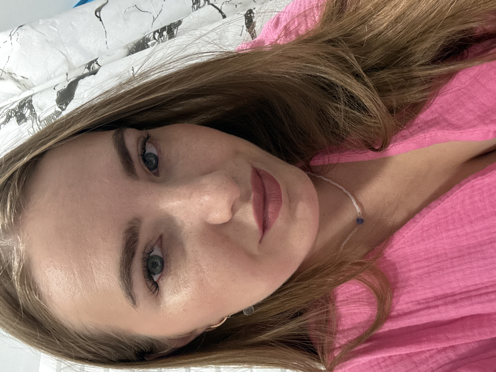 A close-up, eye-level, head-and-shoulders selfie of a young woman with long, light brown hair is captured. She is wearing a bright pink, textured V-neck top and a delicate white beaded necklace with a single blue pendant. A small, silver hoop earring with a gray dangle is visible on her left ear. Her makeup includes mascara, eyeliner, and a matte pink lipstick. The background appears to be a curtain with a black and white birch tree pattern. The lighting suggests an indoor setting. There is no indication of the city of Kazan, Russia.