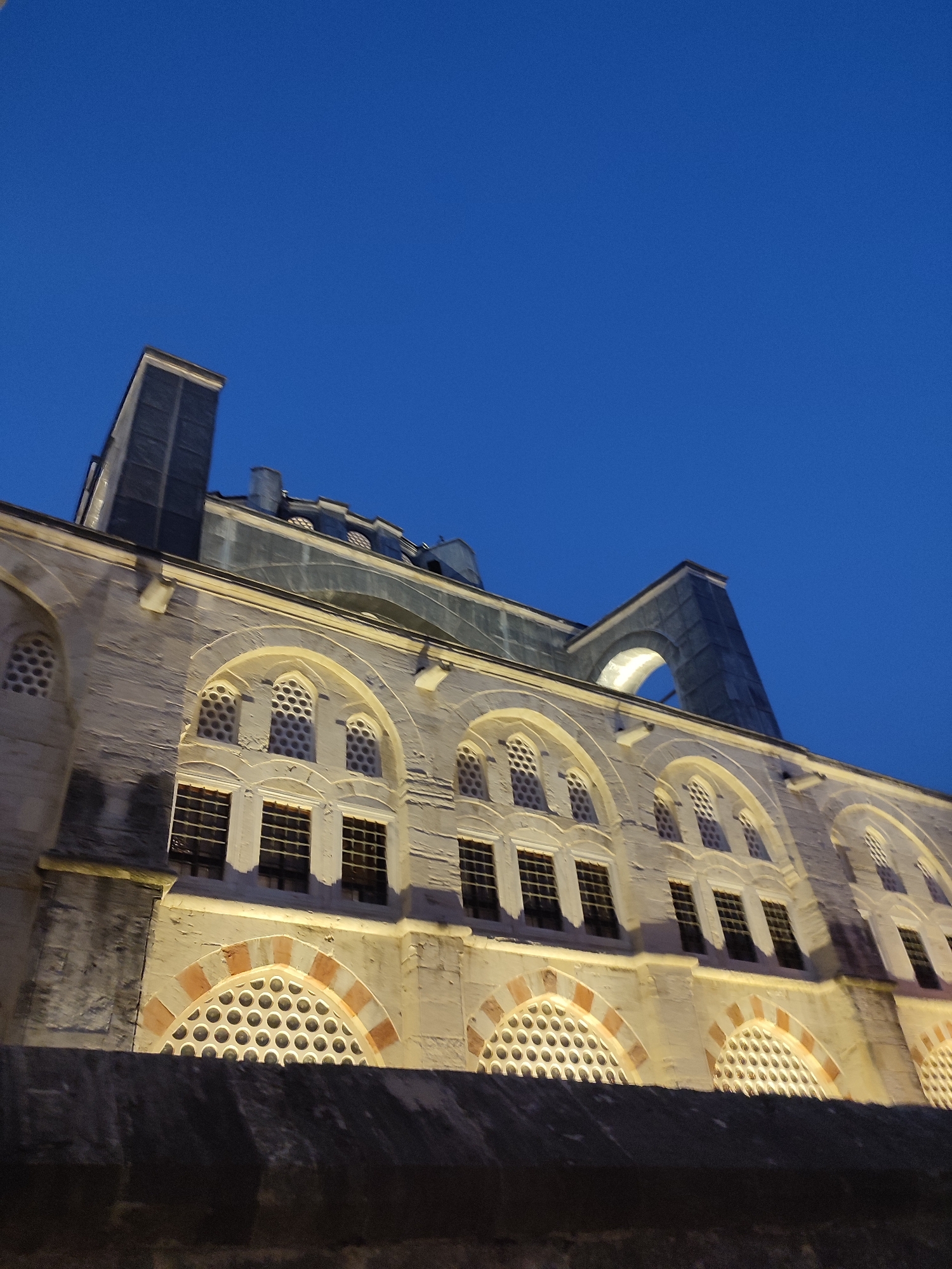 A low-angle, outdoor, night shot captures the facade of a historical building under a deep blue sky. The architecture features light-colored stone with arched windows. Some windows are rectangular with multiple panes and dark frames, while others are smaller and arched with decorative latticework featuring a honeycomb pattern. Red and white striped stone accents frame some of the arched windows. Lighting from below illuminates the lower portions of the building. The upper part of the building shows modern structural elements and a large, grey, arch-like structure against the dark sky. The image is likely from Beyoğlu, Turkey, a district known for its historical architecture and nightlife.