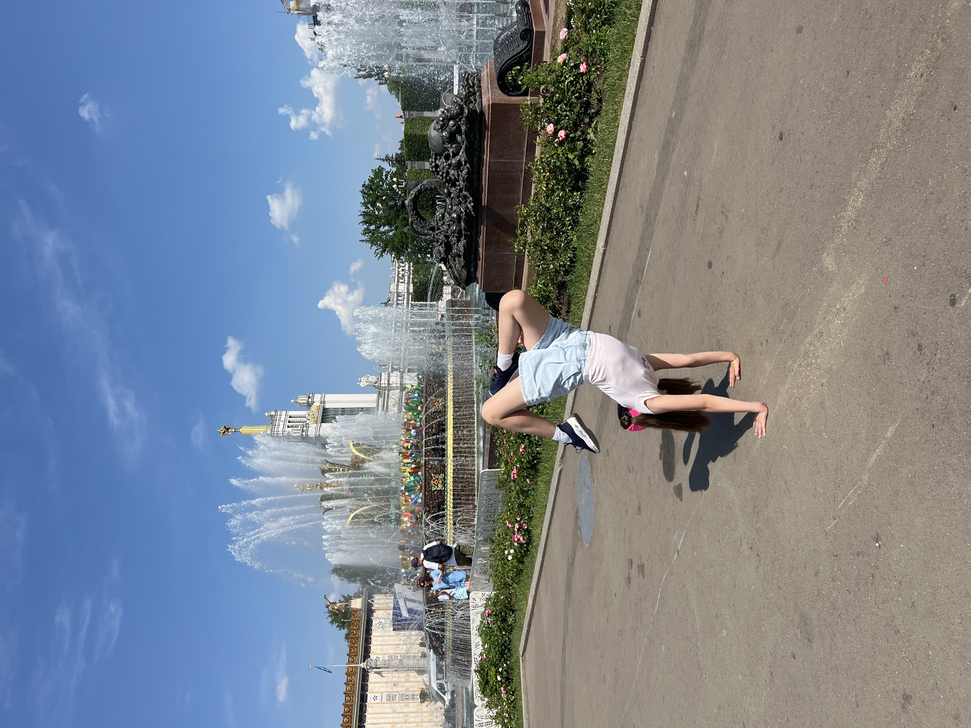 A young girl is performing a handstand in front of a large, ornate fountain with multiple tiers and water spouts. The fountain is surrounded by a paved area and a garden with pink flowers. In the background, there are several buildings, including one with a golden spire topped with a star. The sky is blue with a few scattered clouds. The scene is illuminated by bright sunlight, casting shadows on the ground. The girl is wearing a white t-shirt, denim shorts, and black sneakers. She has her legs in the air, and her hands are on the ground. She is facing away from the camera. Two people are standing near the fountain, observing the water. The location is likely VDNH (Exhibition of Achievements of National Economy) in Moscow, Russia, due to the distinct architecture of the fountain and buildings.