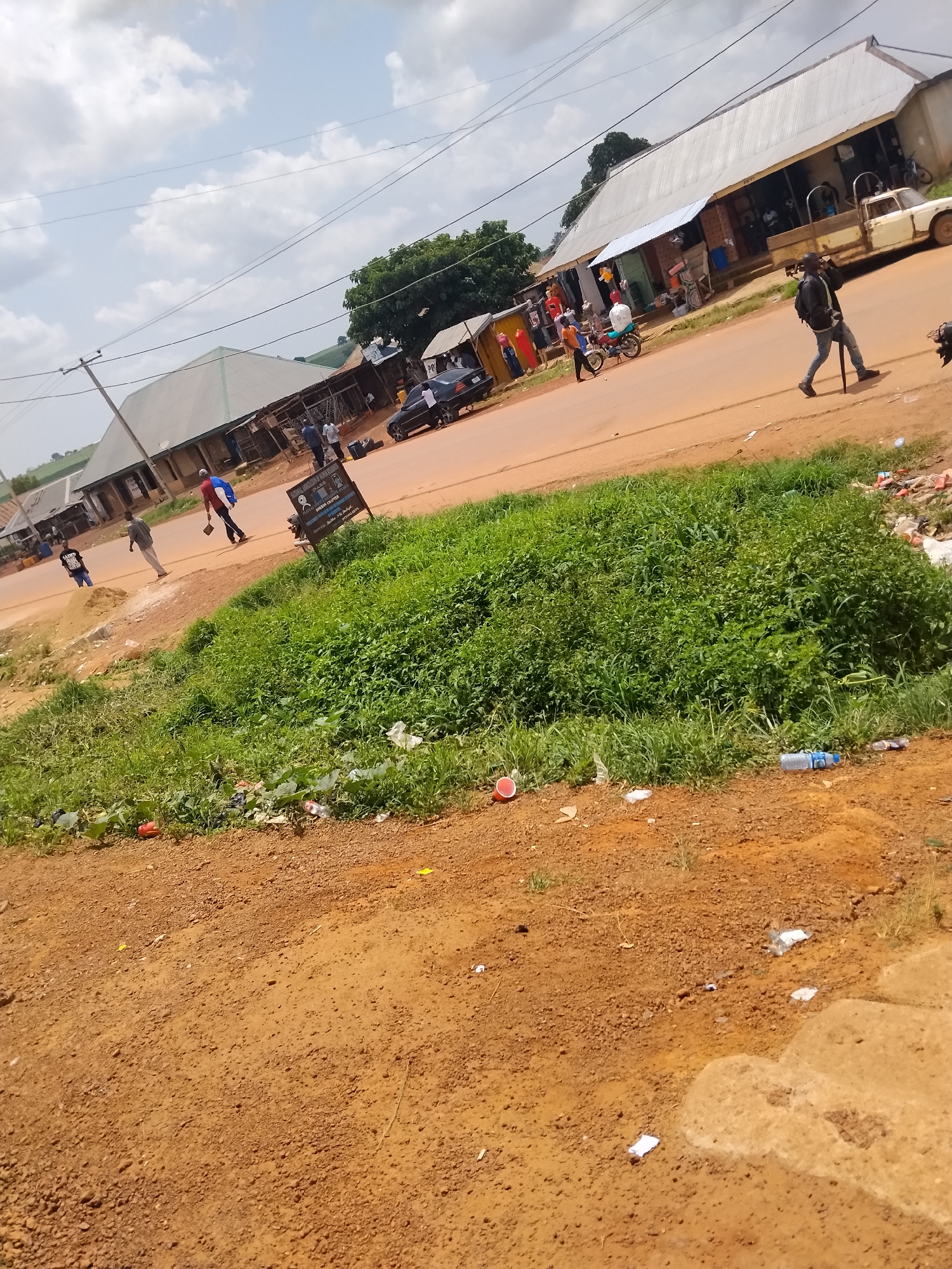 The image depicts a street scene in Durumi, Nigeria, on a cloudy day. A dirt road runs through the center of the frame, with buildings lining the right side. Several people are visible on the road and sidewalks. A man in a black jacket and jeans walks on the road carrying a walking stick, while another person in a red and blue shirt walks towards the left. A white pickup truck is parked in front of one of the buildings, and a white sedan is further down the road. A motorcycle is parked near a stall displaying clothing. The left side of the image shows a large area of dense green vegetation bordering the road. Scattered on the ground are various pieces of litter, including plastic bottles and wrappers. A dark-colored sign with text is positioned near the grassy area. The overall impression is of a typical roadside environment in a Nigerian town, with ongoing activity and evidence of human presence.