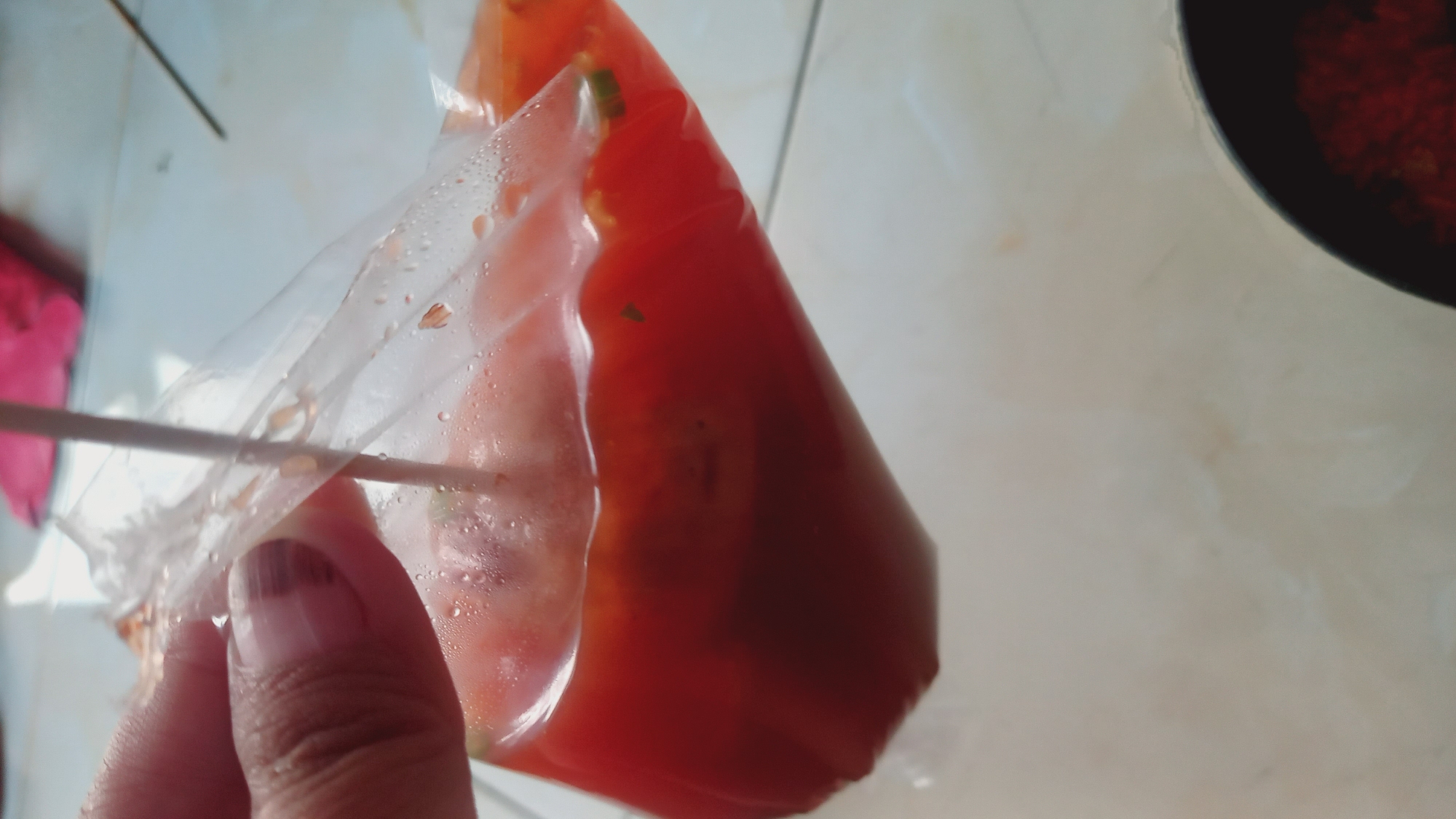 In Leuwisadeng, Indonesia, a close-up shot captures a hand holding a clear plastic bag filled with a red liquid and small food particles. A wooden skewer pierces the bag, and condensation is visible on the plastic's surface. The background suggests an indoor setting with a tiled floor and hints of other objects, including a pink item and a dark bowl containing red food.