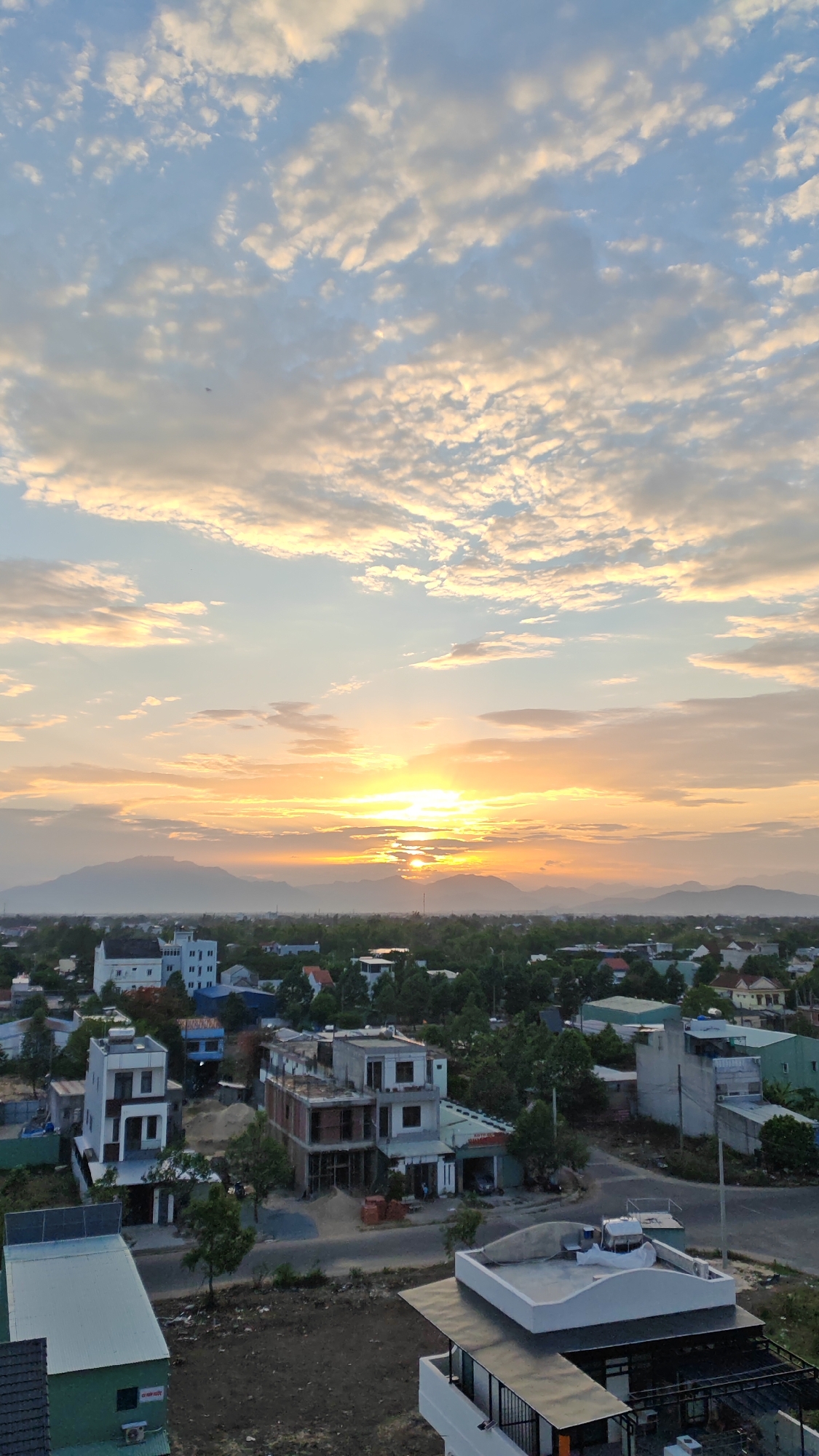 This image captures a panoramic view of a developing urban area at sunset in Đà Nẵng, Vietnam. The sky is dominated by a dramatic display of clouds, illuminated by the setting sun which peeks through them, casting a warm, golden glow across the landscape. In the background, distant mountains are silhouetted against the vibrant sky.
The foreground and midground showcase a dense collection of buildings, ranging from unfinished constructions to completed residential and commercial structures. Many of these buildings are modern, with multiple stories and flat roofs. Construction materials, such as piles of sand and brick, are visible at some sites, indicating ongoing development. Lush green trees are interspersed throughout the urban sprawl, softening the hard edges of the concrete and adding a touch of nature to the scene. Roads and pathways wind through the area, connecting the various buildings.
In the immediate foreground, the roof of a modern building with a white, curved structure is prominent. To the left, another building with a green metal roof is partially visible, with an air conditioning unit mounted on its exterior wall. The overall impression is one of a city experiencing growth and transformation, bathed in the serene beauty of a Vietnamese sunset.