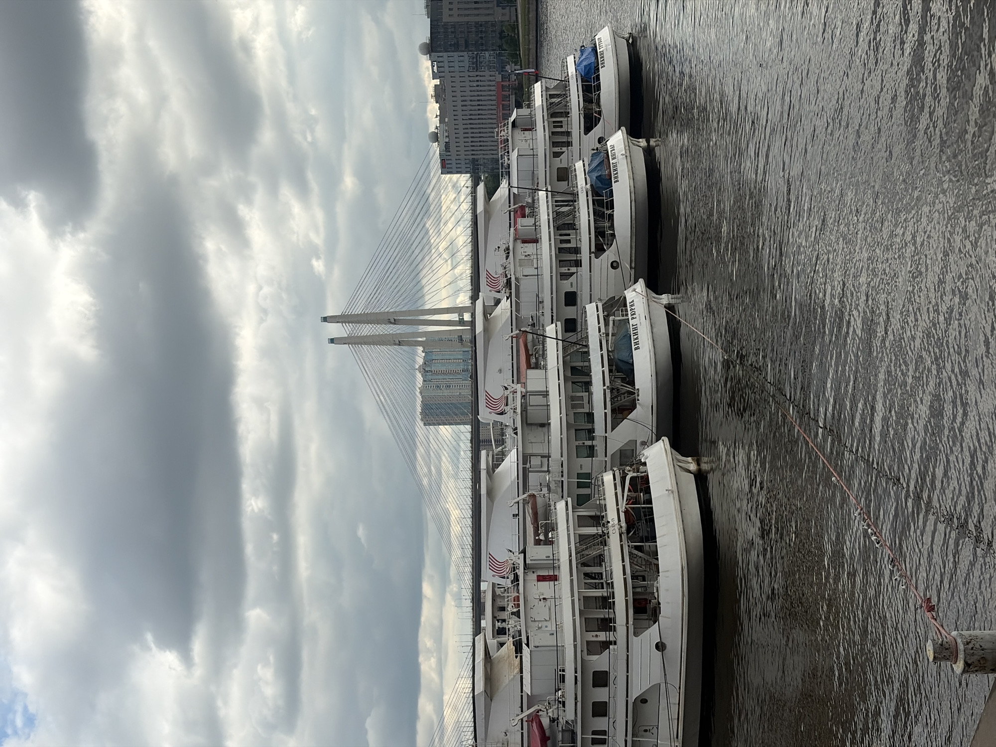 The image depicts a row of docked white passenger boats alongside a body of water, presumably a river or canal, in Saint Petersburg, Russia. In the background, a large cable-stayed bridge is visible, and beyond that, city buildings are discernible. The sky is overcast with heavy clouds. The boats are aligned parallel to each other, with their sides facing the camera. Ropes connect the boats to moorings on the edge of the water.