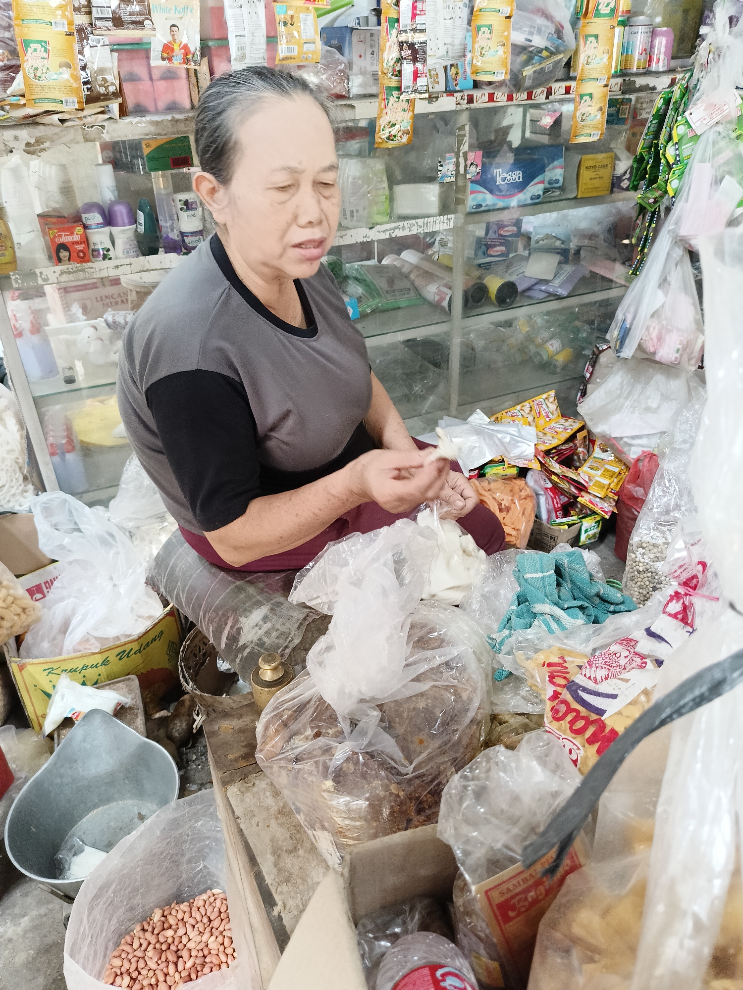 In Barukan, Indonesia, an elderly woman with graying hair is seated behind a stall filled with various goods. She is wearing a gray t-shirt with black sleeves and maroon pants. Her eyes are closed, and her mouth is slightly open as she appears to be speaking or tasting something in her hands, which are holding small white items. The stall is cluttered with a wide assortment of products, including packaged snacks, toiletries, and food items. Shelves behind her are stocked with more merchandise. In the foreground, plastic bags contain nuts, crackers, and other bulk food items. A metal scoop is visible next to a bag of peanuts. The overall environment suggests a small, local convenience store or market stall.