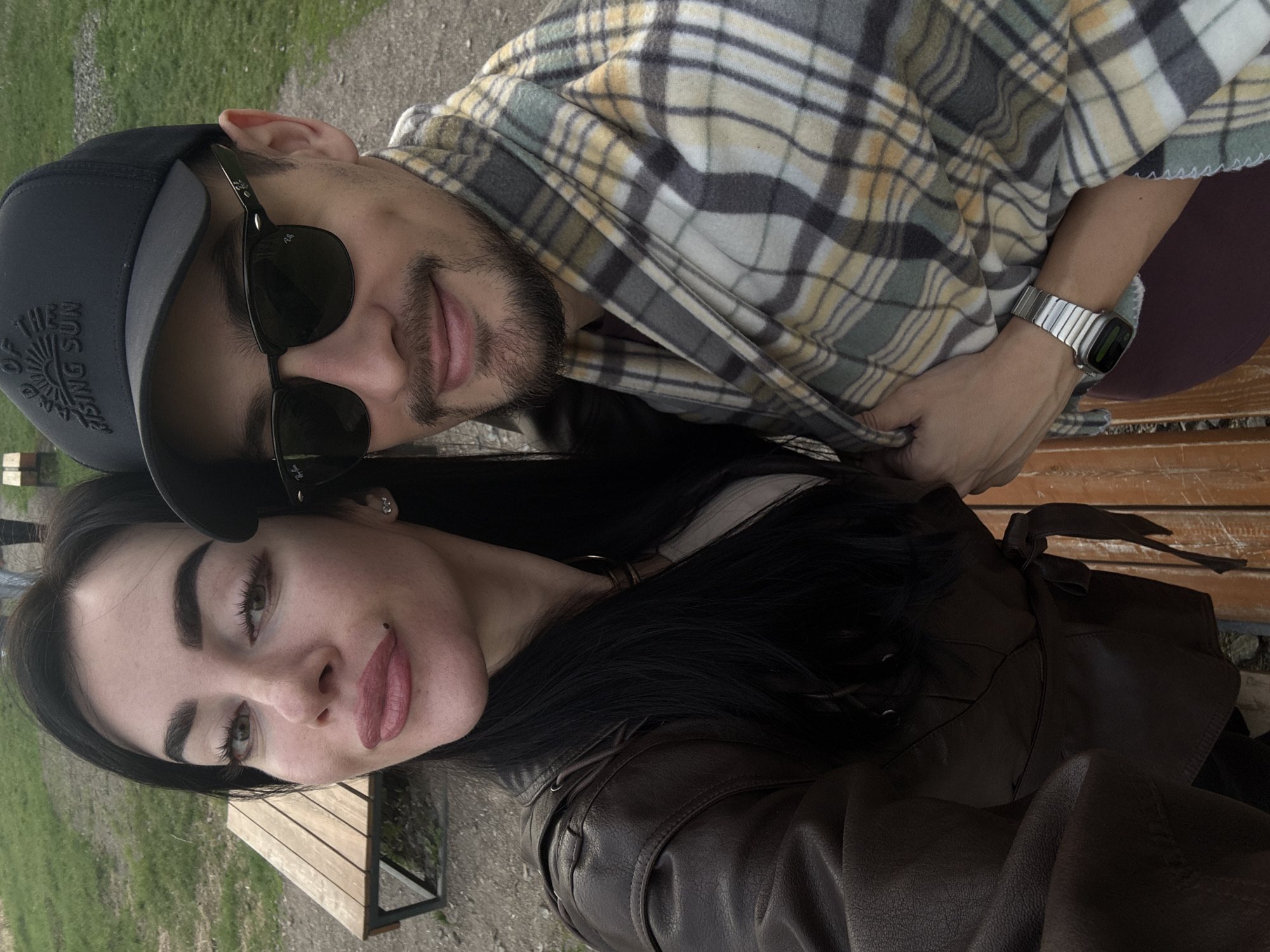 A selfie of a couple. The woman has black hair and wears a dark brown leather jacket. The man is wearing a black cap with the words "Land Of The Rising Sun", sunglasses, and a plaid shirt. A watch is visible on his left wrist. They appear to be sitting on a park bench. The setting appears to be outdoors, with grass visible in the background. The location cannot be determined as Krasnoyarsk, Russia.