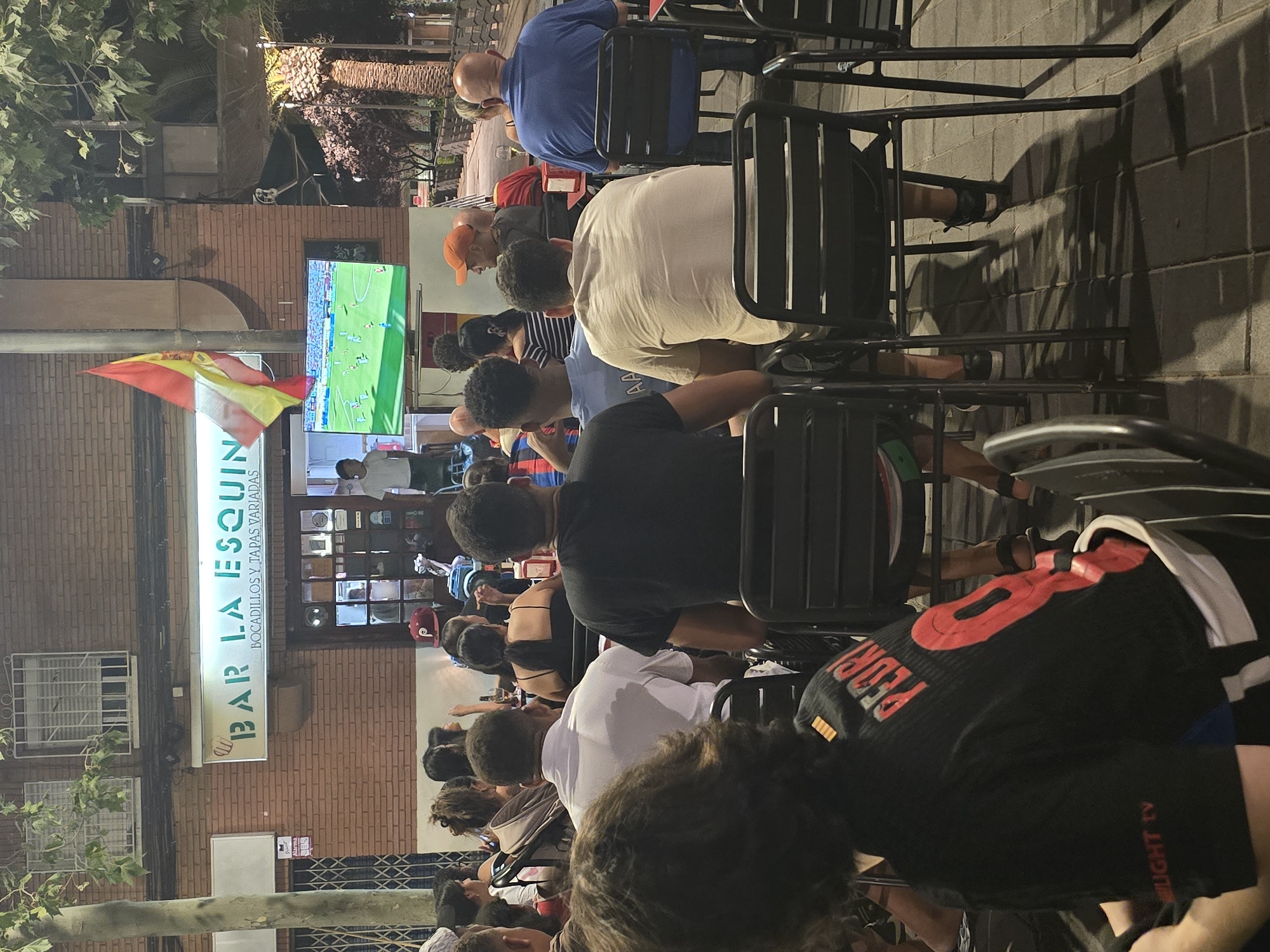 The image captures an outdoor scene at night, likely at a bar or restaurant named "BAR LA ESQUINA" in Cornellà de Llobregat, Spain, as indicated by the signage. A group of people are seated outdoors, facing a television screen displaying a soccer match. The Spanish flag is visible, suggesting a national team game. The setting appears to be a street or plaza with brick buildings, trees, and paved ground. People are casually dressed, some with their backs to the camera, engrossed in watching the game. The atmosphere suggests a communal viewing experience, possibly during a sporting event.