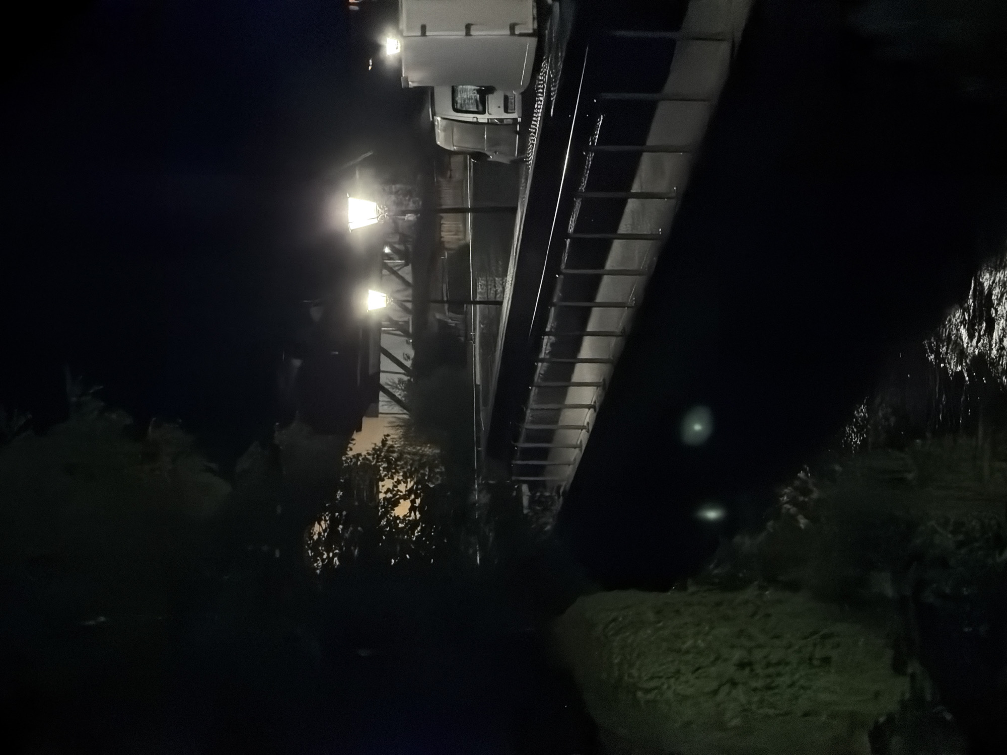 The image depicts a nighttime scene in Garmisch-Partenkirchen, Germany, along the River Partnach. The foreground shows a dark, reflective surface of water, possibly the river, with a textured, mossy bank to the left. A metal railing with vertical posts runs along the right side, bordering a raised platform or walkway.
Beyond the railing, several illuminated streetlights with ornate designs cast a warm glow. In the background, a modern pedestrian bridge or walkway is visible, supported by visible structures. Further to the right, two white utility vehicles, likely parked, are partially illuminated by the streetlights, one appearing to be a van and the other a larger, box-shaped vehicle. The overall atmosphere is calm and dimly lit, emphasizing the architectural elements and the artificial lighting against the dark night sky.