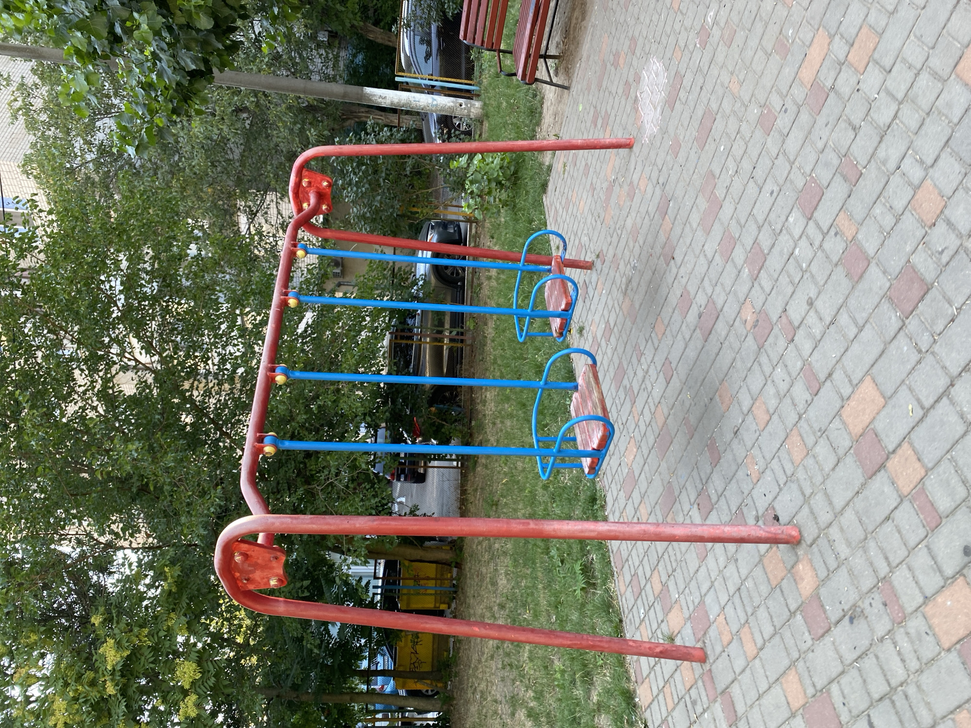 An outdoor scene in Chornomorsk, Ukraine, features a red metal swing set with two blue swings, each with a wooden seat and blue frame. The swing set is situated on a paved area of interlocking bricks in red and grey hues. Behind the swing set, a patch of green grass is visible, with trees providing a backdrop. Several parked cars are present in the background, along with a portion of a multi-story building. To the right of the swing set, a red park bench is positioned on the paved surface. The overall environment suggests a public recreational area within a residential or urban setting.