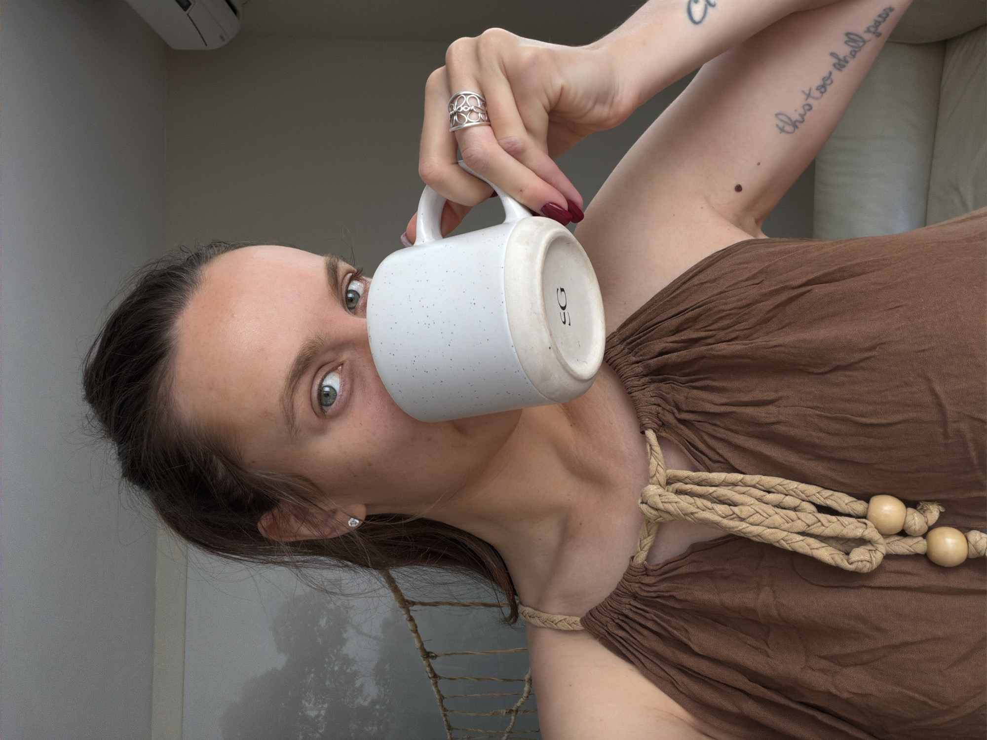 A woman is shown at a tilted angle, holding a white mug with dark speckles and a tan bottom. The mug is positioned against her face, obscuring part of it. Her right hand, adorned with a large silver ring and dark red nail polish, grips the mug's handle. Visible parts of her face reveal light skin and piercing green eyes. She has dark hair pulled back and wears a small, circular earring. Her right arm is raised, revealing tattoos on her forearm. She wears a brown, flowing top with a tan rope necklace. The background suggests a simple room with a partial view of a white couch and an air conditioning unit on the wall. Outside the window is a tree. The photograph appears to be taken indoors in Budva, Montenegro.