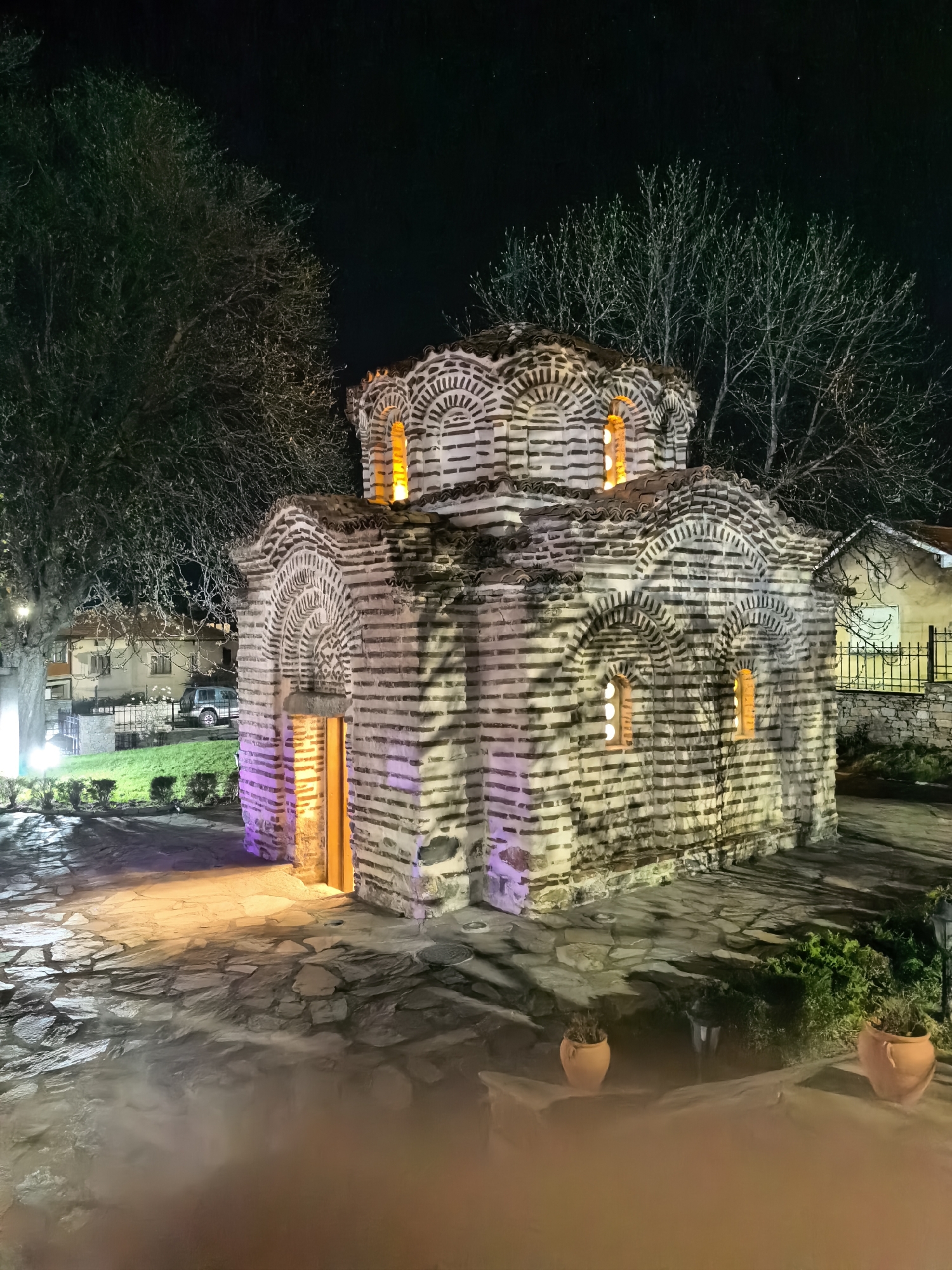 The image captures the Church of Sveti Pantelejmon at night in Ohrid, North Macedonia. The Byzantine-style church, made of stone and brick, features illuminated windows and a doorway. It stands on a stone-paved area surrounded by trees and glimpses of nearby buildings. Potted plants are visible in the foreground.