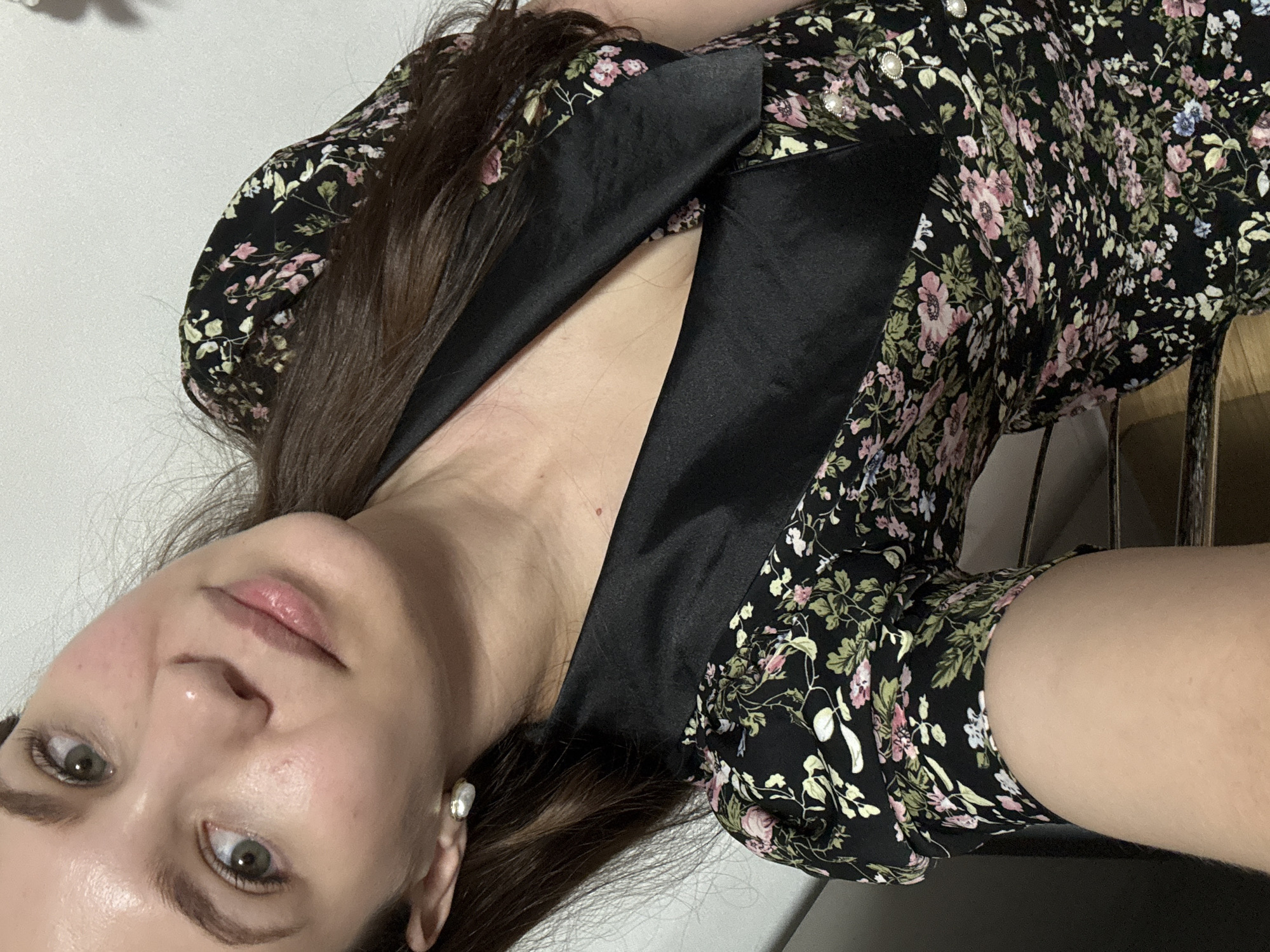 A young woman with light skin and brown hair is shown in a close-up portrait. She is wearing a black dress with a floral print featuring pink and green flowers. The dress has a black satin collar and pearl-like buttons. She has green eyes and is wearing pearl earrings. The background is a plain white wall. The image was taken in Chaikovsky, Russia.