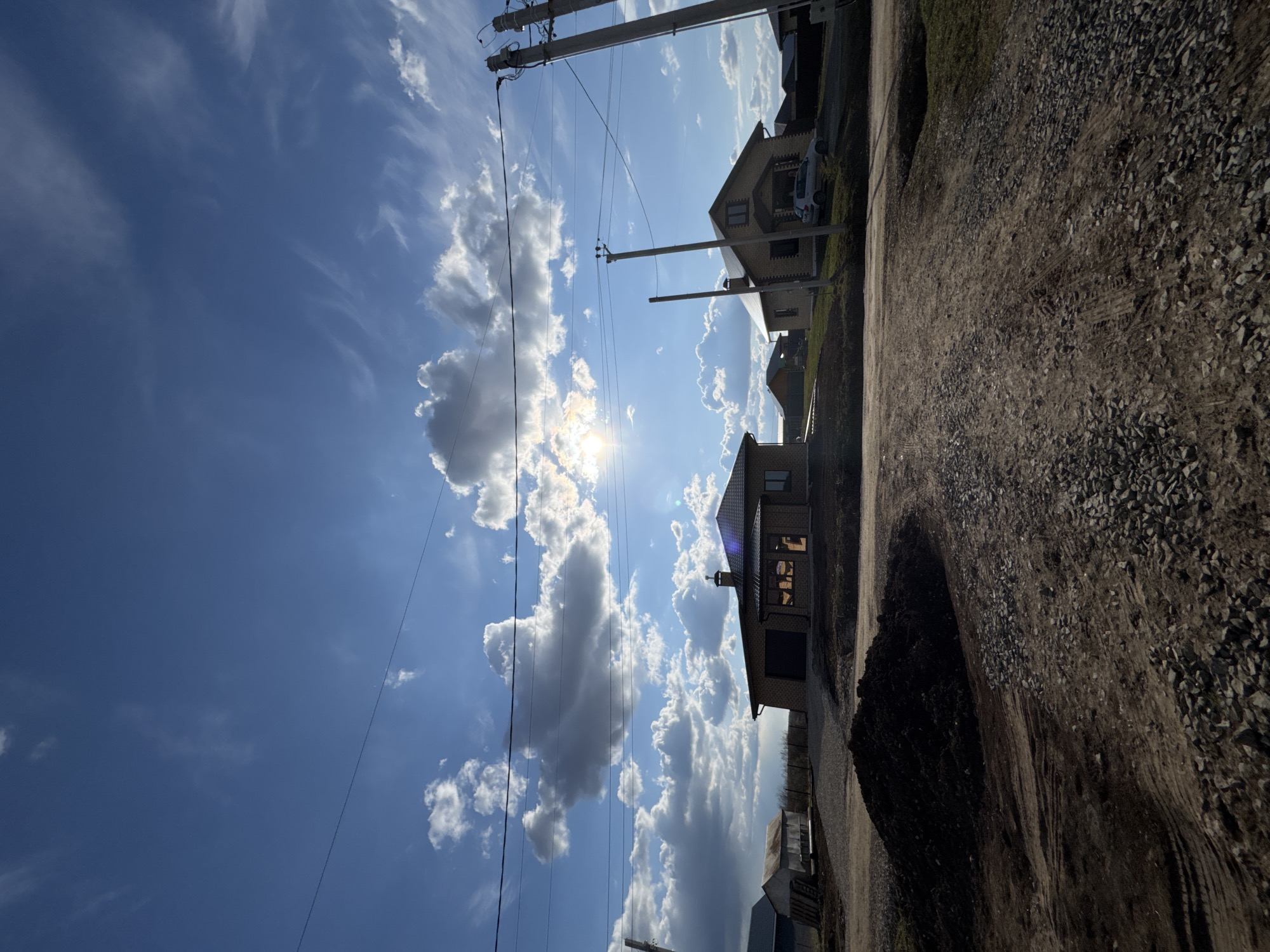 The image depicts a view in the city of Gornyi (Dvurechenskoe S/P), Russia. The scene is outdoors on a sunny day with blue sky and white clouds. In the foreground, there is a gravel road. On the right side, there are one or two houses, and some utility poles/wires. The sun is shining brightly, partially obscured by the clouds.