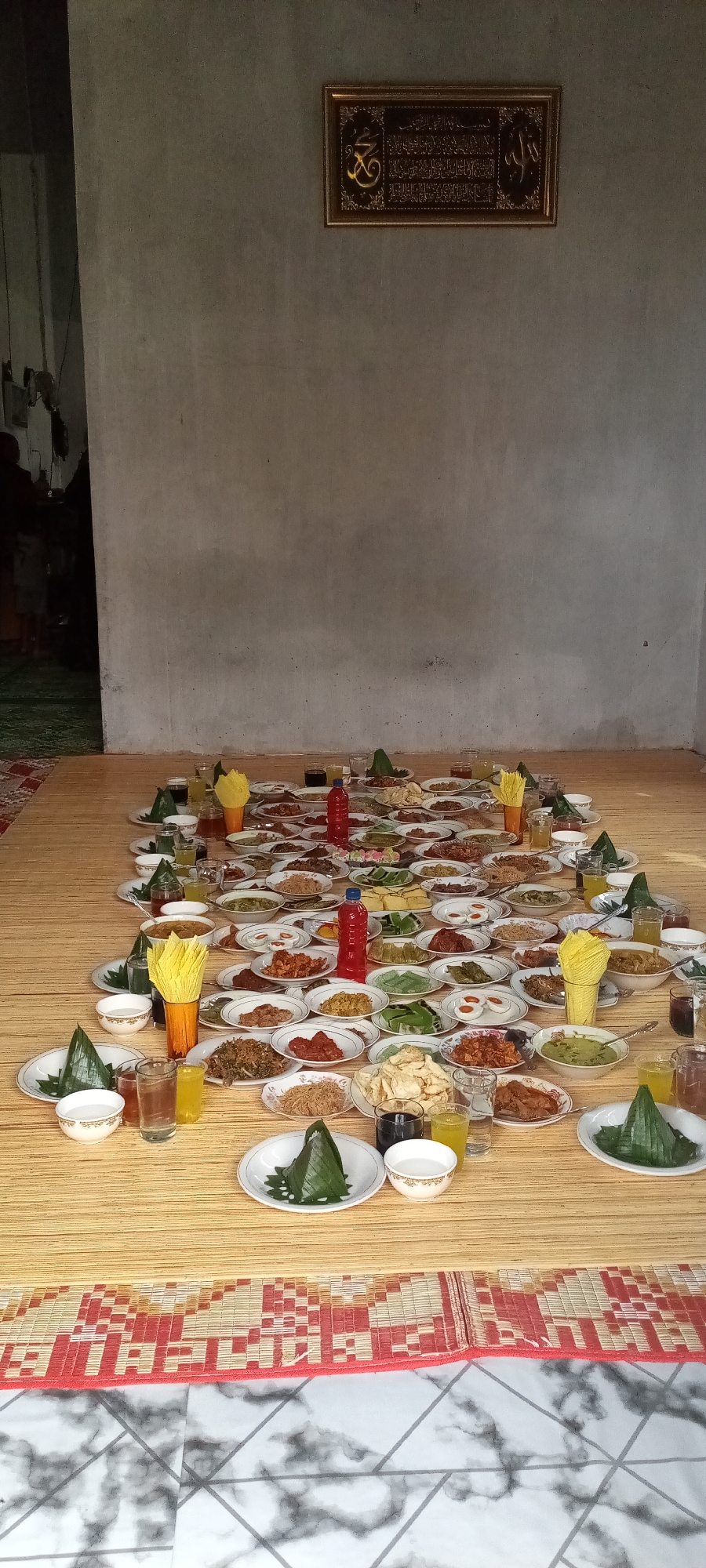 This image captures an indoor scene in Lhokseumawe, Indonesia, depicting a traditional communal meal setup. A vast array of Indonesian dishes, served on numerous small, round white plates, are meticulously arranged across a large, light brown woven mat that covers the floor. The plates contain various preparations, including meats, vegetables, crispy items, and what appear to be savory snacks or desserts. Several small, cone-shaped items wrapped in green banana leaves, likely containing rice or a similar food, are also interspersed among the plates.
Numerous clear glasses containing liquids of varying colors (clear, yellow, and dark) are positioned throughout the food spread, along with a few small white bowls, possibly for sauces or additional beverages. Two prominent red plastic bottles, likely containing drinks or condiments, are visible. Stacks of yellow paper napkins are presented in orange or brown holders at several points.
The room features plain, light grey concrete walls with a visible rough texture. Centered on the upper wall is a framed rectangular artwork with a golden frame. The artwork contains intricate golden Arabic calligraphy on a dark background, likely a religious inscription. To the far left, a dark doorway reveals a glimpse of an adjacent room with a green patterned mat on the floor and a dark, indistinct figure partially visible within the shadows. The floor beneath the main woven mat is partially visible at the bottom of the image, showing a white and grey marbled tile pattern with geometric lines. A red and white patterned mat acts as a border between the woven mat and the tiled floor. The overall lighting is even and ambient. No individuals are actively engaged with the food, suggesting the arrangement is complete or awaiting guests.