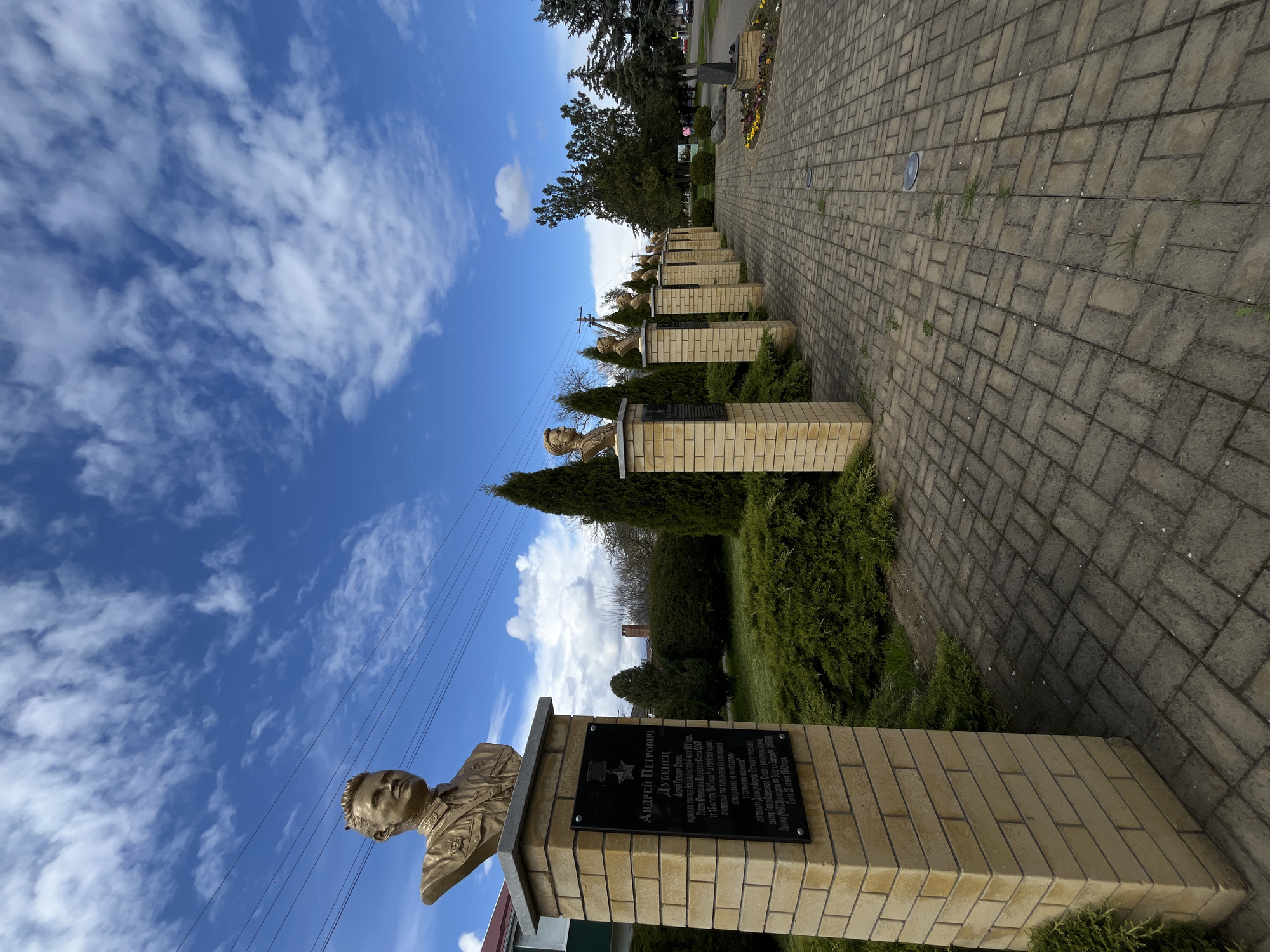 The image depicts a row of bronze busts mounted on brick pedestals in what appears to be a memorial or cemetery in Novopokrovskaya, Russia. The busts are of military figures, likely soldiers or officers, as indicated by their military attire. Each pedestal has a black plaque bearing inscriptions in Russian. The plaques identify the individuals as Andrey Petrovich Dubenets. The background features trees, shrubbery, and a paved pathway. The sky above is blue with scattered clouds. Power lines are visible. The overall scene conveys a sense of solemnity and remembrance.