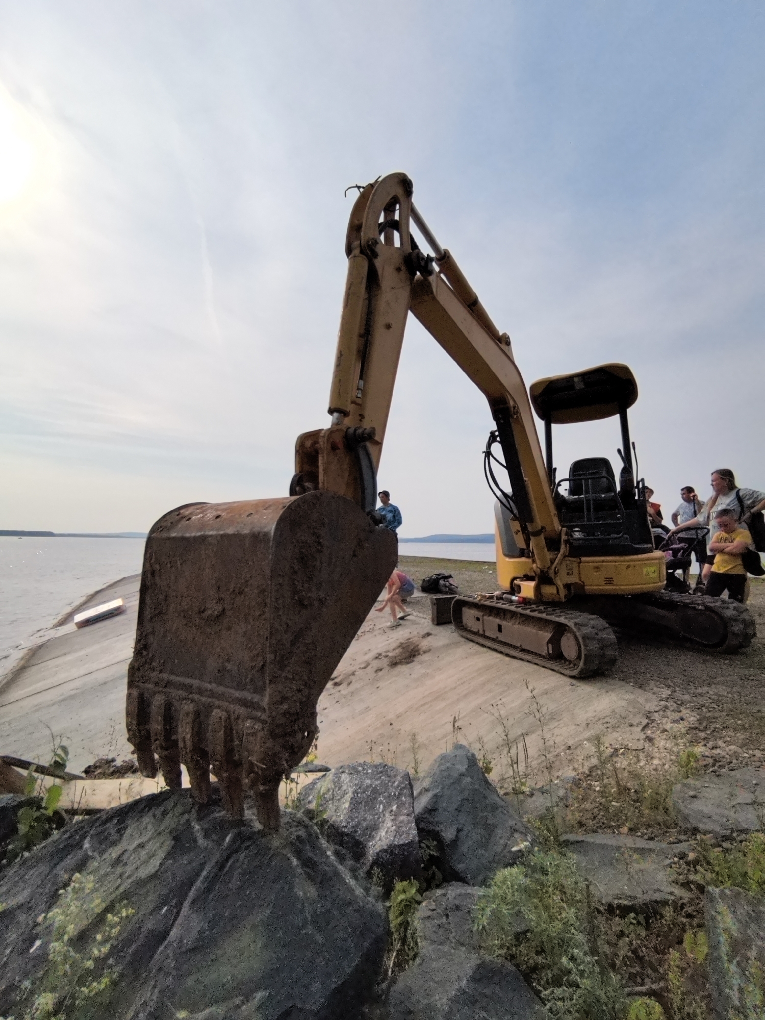 An excavator is positioned on a dirt embankment near a body of water. Several people are gathered around the excavator, some appearing to be watching or interacting with it. The excavator has a large bucket that is partially obscured by rocks in the foreground. The sky is partly cloudy.