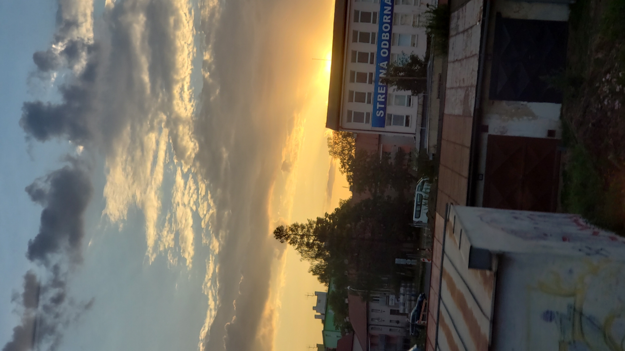 The image depicts a view of Poprad, Slovakia, during sunset. The sky is a blend of blue and orange hues, with scattered clouds. In the middle ground, a building with the text "STREDNÁ ODBORNÁ" is visible. Several trees are scattered throughout the scene, and in the foreground, there are garages with rusty roofs and graffiti on the walls. A van and a car are parked on a street behind the garages. The overall impression is of an urban landscape illuminated by the warm glow of the setting sun.