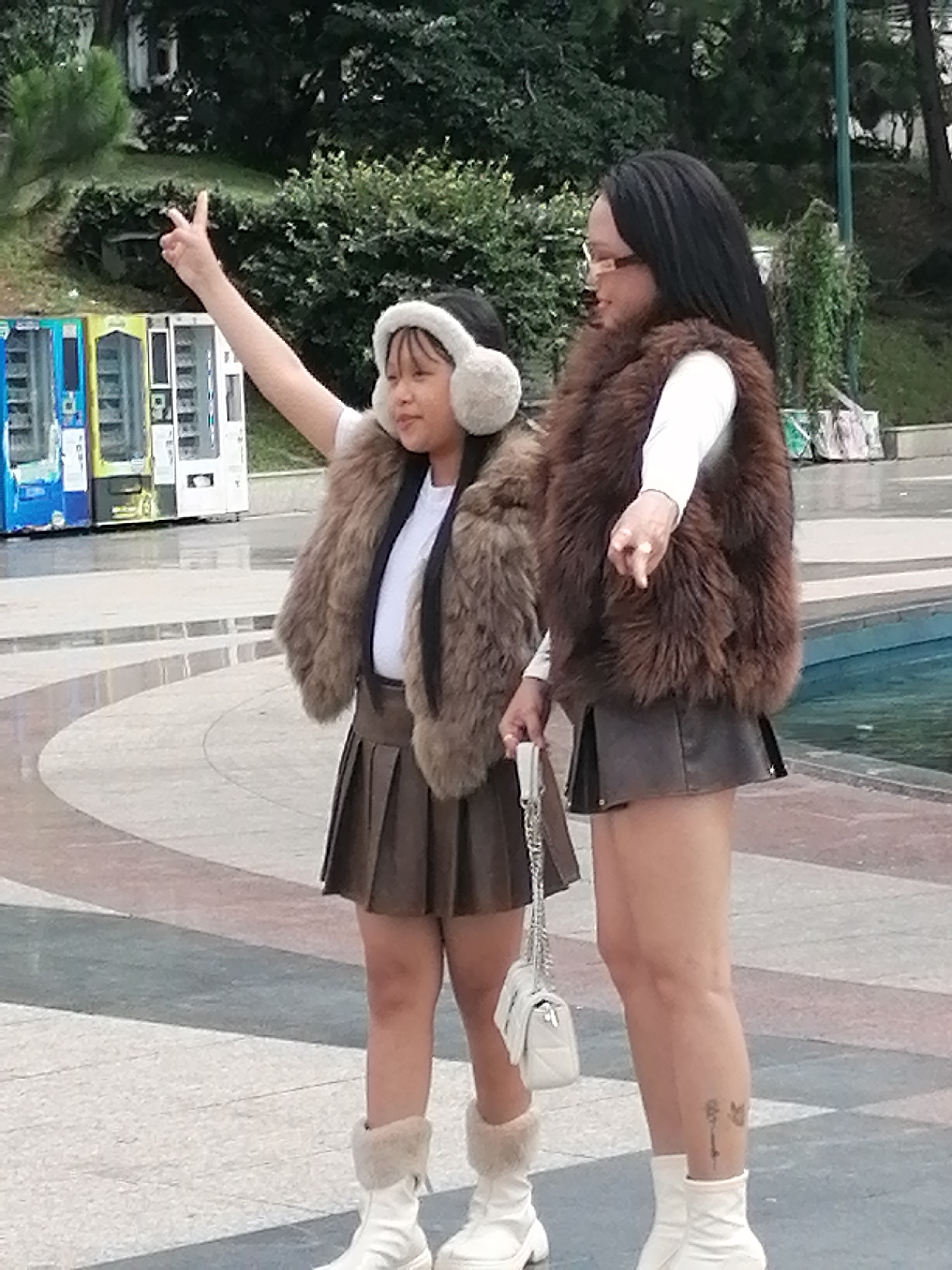 The image depicts two individuals, a younger person and an older person, standing outdoors on a paved surface with a water feature to their right. The younger person, on the left, is wearing a brown faux fur vest over a white shirt and a brown pleated skirt, with white boots and fluffy earmuffs. They are raising their left hand with two fingers extended in a peace sign. The older person, on the right, is wearing a similar brown faux fur vest over a white long-sleeved shirt and a dark brown leather skirt, paired with white boots. They are pointing their right index finger forward. Both individuals are facing towards the left side of the frame, with the younger person looking slightly towards the viewer. In the background, there are several vending machines, lush green trees, and a water fountain. The overall setting suggests an urban park or public space in Da Lat, Vietnam.