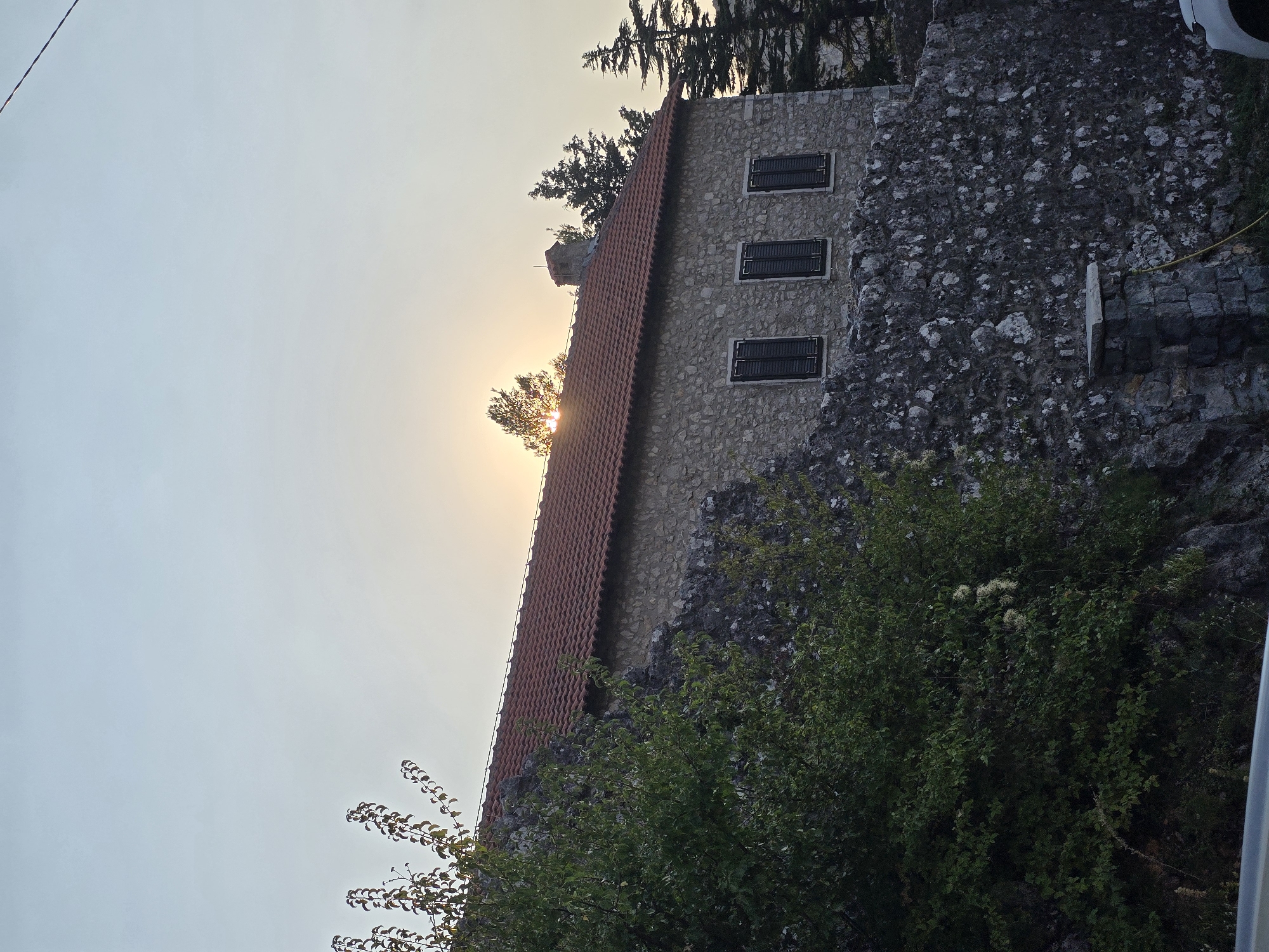 The image shows a stone building with a red-tiled roof, situated in Medun, Montenegro. The building has three dark-shuttered windows. In front of the building is a rough stone wall, with a dense green tree to the left. The sun is setting behind some trees on the right side of the frame, casting a warm glow and creating a lens flare. The sky is a pale, hazy blue. A small portion of a white car is visible in the bottom right corner.