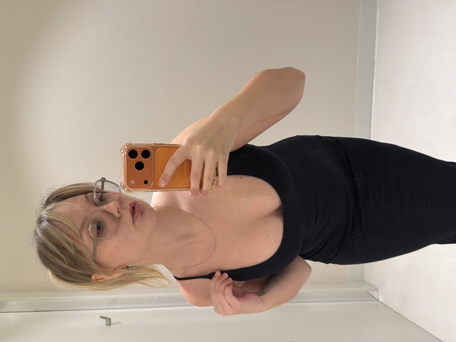 The image depicts a young adult female captured in a mirror selfie within what appears to be a fitting room, described as being located in Tel-Aviv, Israel. The female has light skin, blonde hair styled in a ponytail with bangs, and wears clear-framed glasses, small dark earrings, and a thin gold-colored chain necklace. She is dressed in a form-fitting, scoop-neck, black midi-length dress with thin spaghetti straps. Visible tan lines are present on her upper chest and shoulders, indicating prior sun exposure. Her right hand holds an orange-cased smartphone, equipped with a triple-lens camera module, pointed towards the mirror for the selfie. Her left hand is touching the left strap of her dress. The background consists of plain, light-colored walls. A white door frame and a chrome wall hook are visible on the left side of the frame. The floor is light-colored, featuring a darker baseboard.