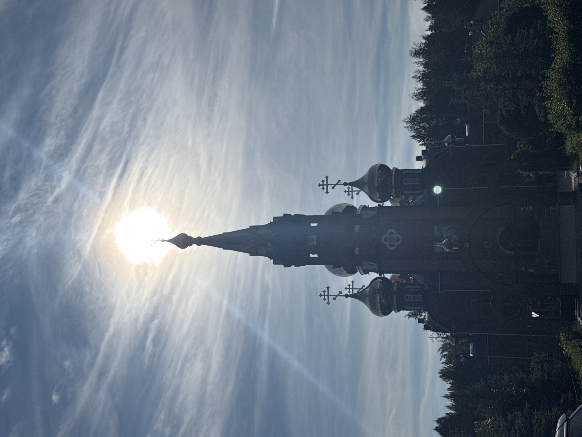 An outdoor shot captures a striking view of an Orthodox church silhouetted against a bright, cloudy sky. The image is oriented vertically (portrait mode), with the church dominating the right side of the frame and the sky filling the left and upper portions.
The main subject is a large, dark church building featuring a prominent, tall central spire that tapers upwards to a point, topped with a cross. Around the main structure, several golden onion domes are visible, each also crowned with an intricate Orthodox cross. The church's facade is heavily shadowed due to the intense backlighting, making architectural details difficult to discern, but some decorative patterns and window shapes can be faintly made out.
To the left of the church's main spire, the sun is directly visible as a brilliant, almost white orb, causing a strong glare and lens flare effects that extend across the sky. The sky itself is light blue, filled with delicate, wispy cirrus clouds that stretch across the frame, creating dramatic streaks and patterns.
Behind and to the far right of the church, a dense line of dark green trees forms a natural backdrop. No people or visible text are present in the image. The scene depicts a bright daytime, likely late morning or afternoon, with clear weather conditions allowing for brilliant sunlight. The architectural style strongly suggests an Eastern Orthodox church, fitting the location context of Chaikovsky, Russia.