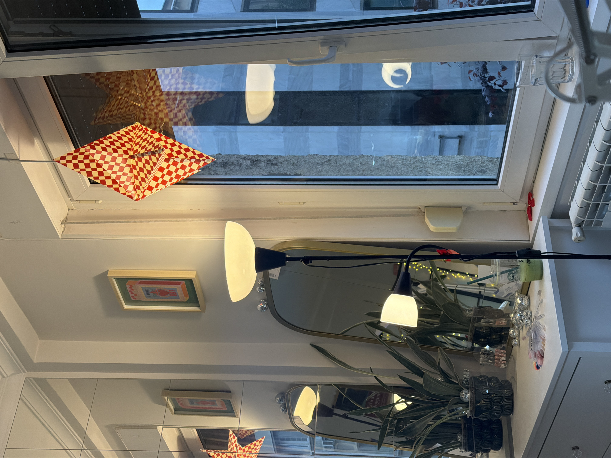 The image shows an interior view looking out through an open window. A red and white checkered lantern is hanging from the window frame, with a larger star-shaped lantern behind it. Outside, the view is of buildings with visible windows and architectural details. Below the window, there is a scene with a lamp, a framed picture, a mirror, and a plant in a black pot. A Starbucks cup sits on the window sill alongside some decorative balls and feathers. The photo seems to have been taken indoors, with the camera tilted upwards. The location is Belgrade, Serbia, based on visual clues.