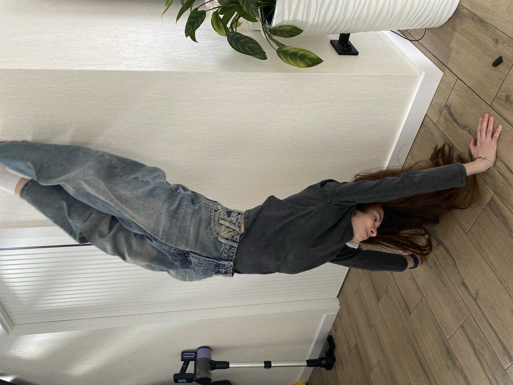 A woman lies on a wooden floor in a room. She is wearing jeans and a long-sleeved shirt. Her hair is spread out on the floor. One hand is outstretched towards the top right of the image. A plant in a white pot is visible in the upper center. A vacuum cleaner is near the bottom. The room has white walls. The location is Votkinsk, Russia.