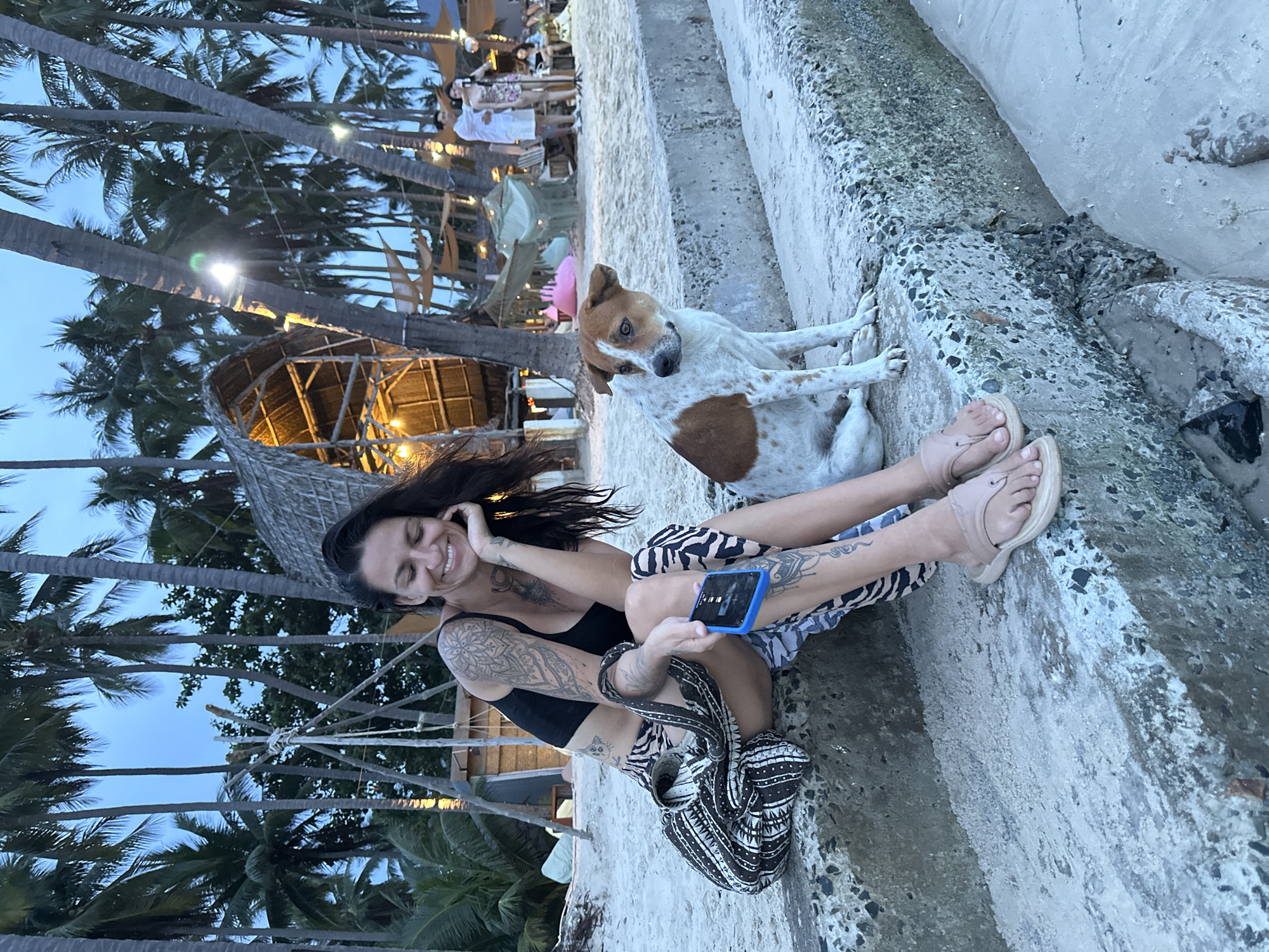 A woman with dark hair and extensive tattoos is sitting on concrete steps on a beach. She is wearing a black bikini top and patterned shorts. She is smiling and holding a blue phone. A dog with brown and white markings is sitting next to her, also on the steps. In the background, there are palm trees, thatched-roof structures, and beach umbrellas. The scene appears to be a tropical beach resort. The lighting suggests it is either early morning or late afternoon. The sky is a pale blue. The overall mood is relaxed and casual.
