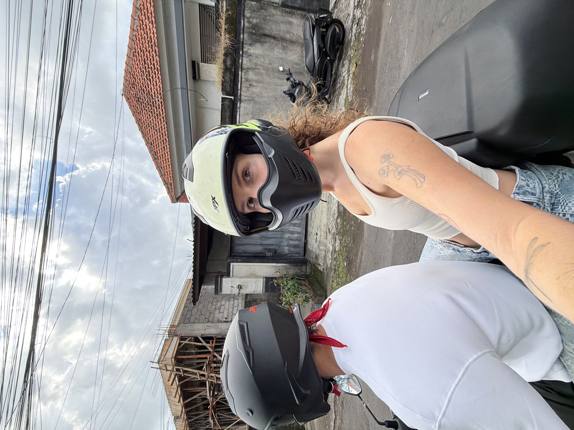 Two individuals are riding a scooter on a paved road in North Kuta, Indonesia. The person in front, on the right, is a woman wearing a light-colored helmet with a black face mask and a white tank top. She has curly brown hair and a tattoo of mushrooms on her right arm. The person behind, on the left, is wearing a black helmet and a white t-shirt, with a red bandana tied around their neck. Both are looking away from the camera. In the background, a black scooter is parked. To the left, a building under construction is visible, with scaffolding and concrete structures. To the right, a residential building with a red tile roof and a grey concrete wall is present. Overhead, power lines crisscross the cloudy sky. The overall scene depicts a typical street in a developing area.
