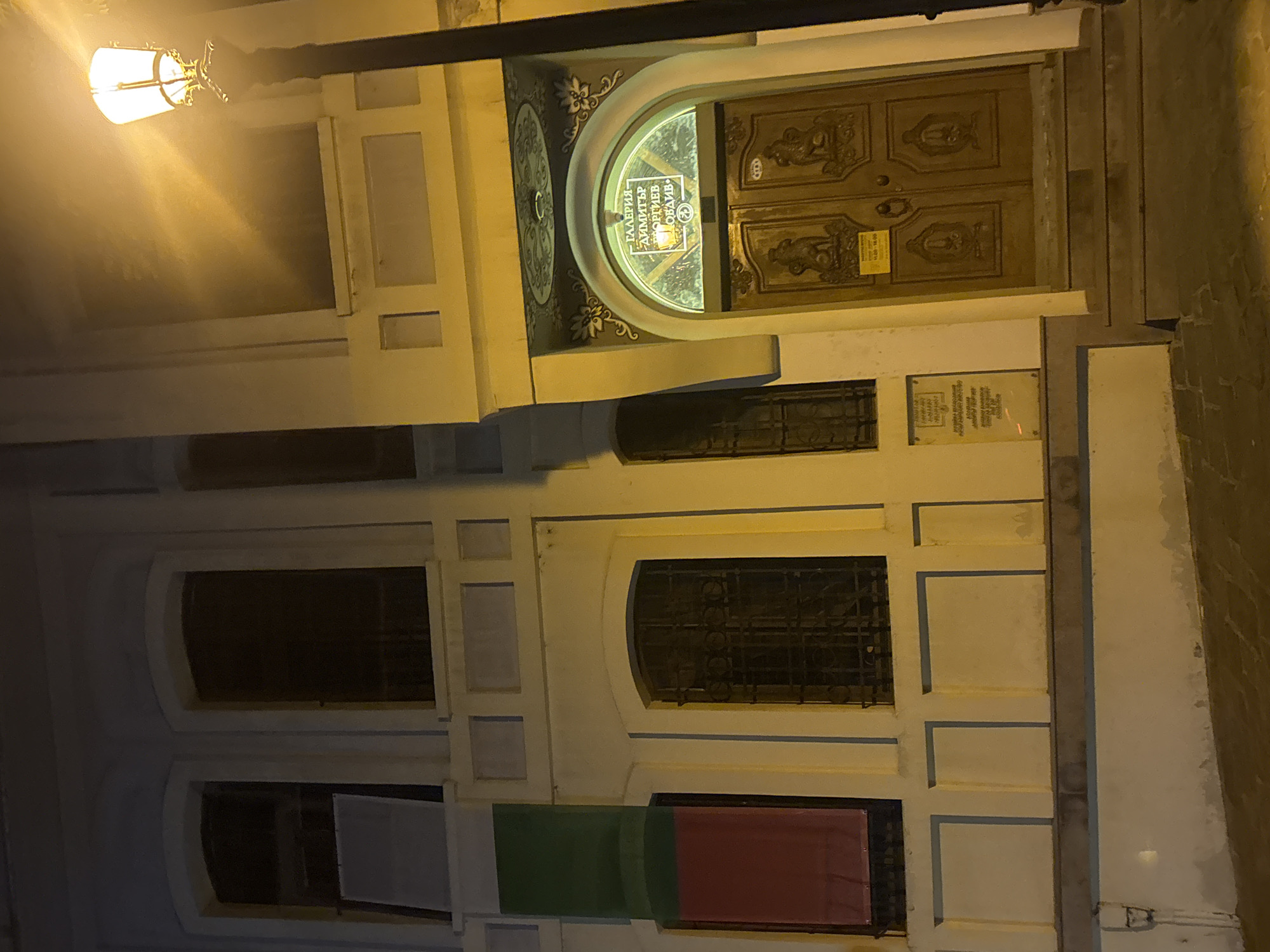 This image depicts the entrance to the Dimitar Georgiev Gallery in Plovdiv, Bulgaria, at night. The building's facade is painted in light colors with darker accents around the windows and entrance. A streetlamp with a warm, yellow glow illuminates the right side of the scene, casting long shadows.
The gallery entrance features an arched doorway with ornate golden double doors. Above the doors, a semi-circular sign displays text in what appears to be Cyrillic script, illuminated from within. Decorative floral motifs are visible above the archway. The doors themselves are intricately carved with what seem to be lion or sphinx-like figures and other decorative elements. A plaque with text is affixed to the wall to the left of the entrance, near a window with ornate metal bars.
To the left of the gallery entrance, the building has several windows, some covered with what appears to be fabric. One window on the left displays a banner with horizontal stripes of green and red fabric. The overall scene suggests a quiet, possibly historic, part of the city illuminated by artificial light. No people are visible in the image.