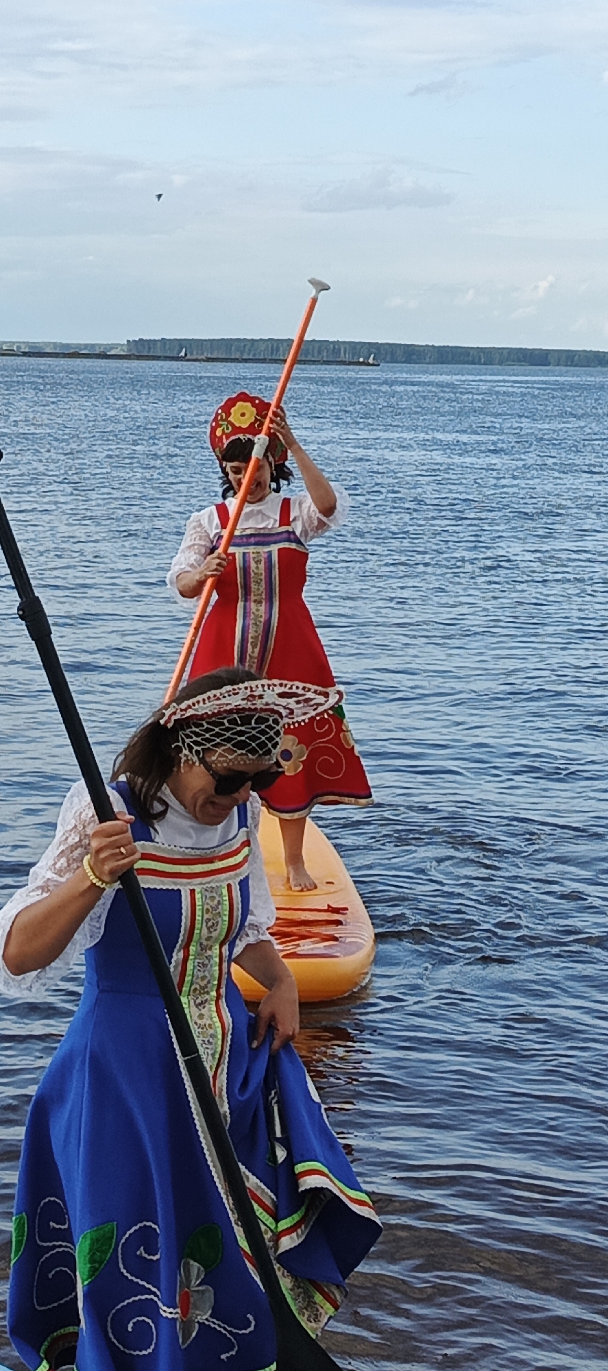 Two women in traditional Russian attire are paddleboarding on a body of water, possibly the Kama River, near the city of Chaikovsky, Russia. The woman in the foreground wears a blue dress with white lace sleeves and a decorated bodice, and she holds a paddle. The woman behind her wears a red dress with similar decorative elements and a traditional headdress. Both are on an orange paddleboard. The water is calm, and the sky is overcast. The background shows a distant shoreline with trees and some structures.