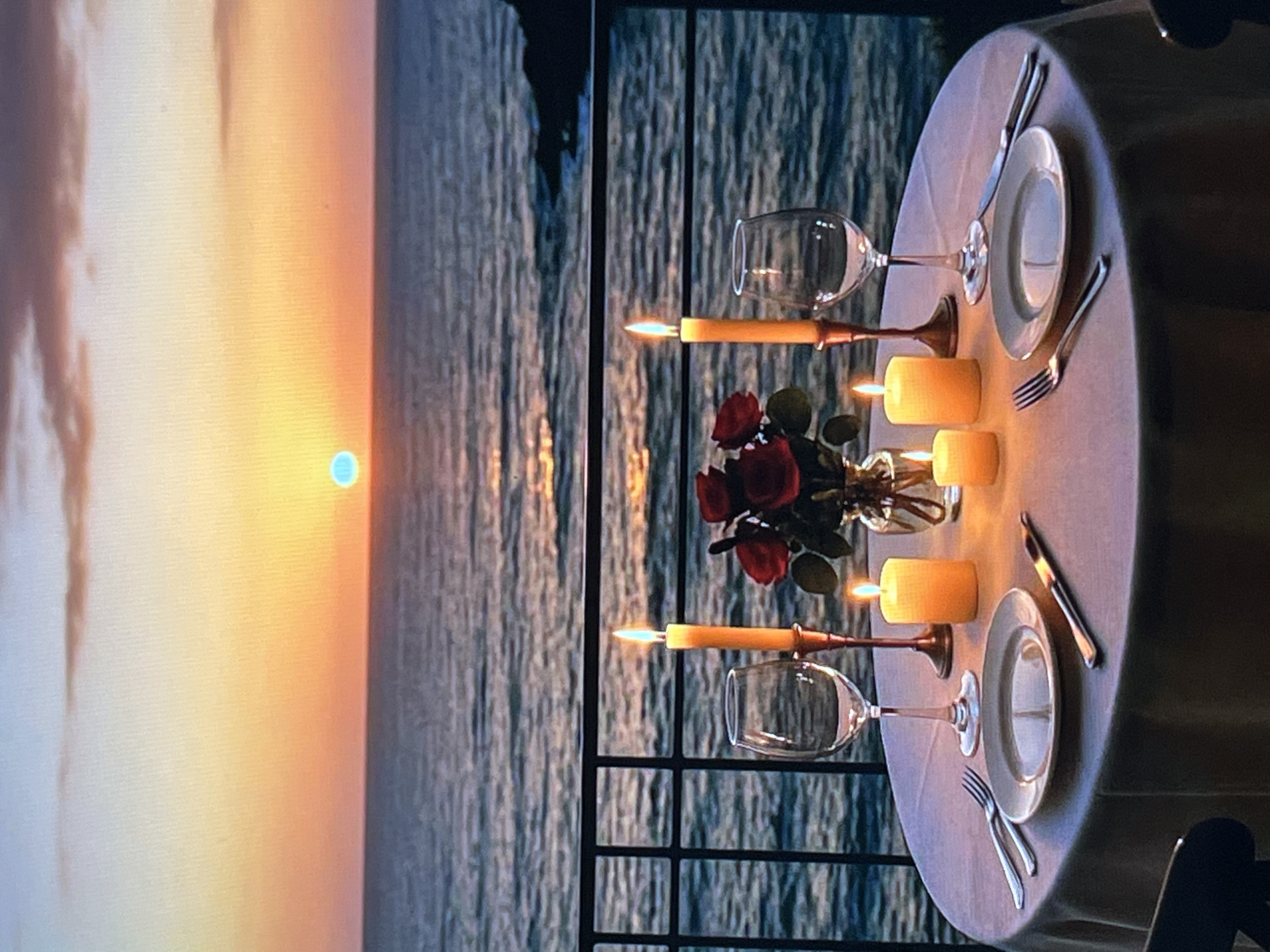 A romantic dinner for two is set on a balcony overlooking the ocean at sunset. The table is covered with a light-colored tablecloth and set with two place settings, each including a plate, silverware, and a wine glass. Two candles are lit, casting a warm glow on the table. In the center of the table, a small vase holds a bouquet of red roses. The sunset casts a warm orange and yellow hue across the sky and reflects on the water. The mood is serene and intimate, perfect for a special occasion.