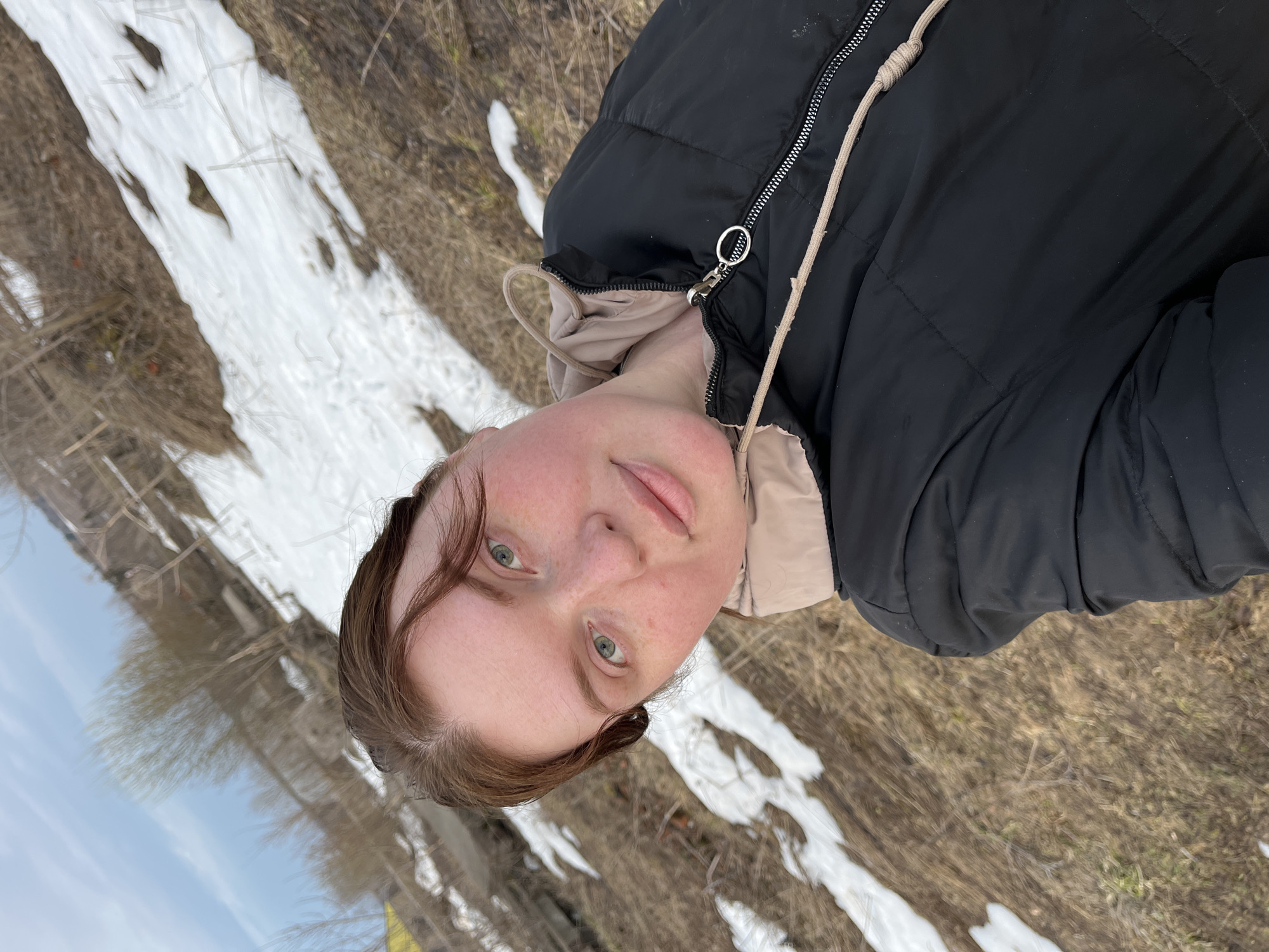 A young woman is the main subject of the image, positioned in the foreground and slightly off-center. She has fair skin, light eyes, and brown hair styled with a few strands framing her face. She wears a black puffy jacket over a light-colored hooded sweatshirt. The background suggests an outdoor setting in the town of Павловка, Russia, as patches of melting snow cover the ground, mixed with dry grass and vegetation. The sky is faintly visible in the top left corner.