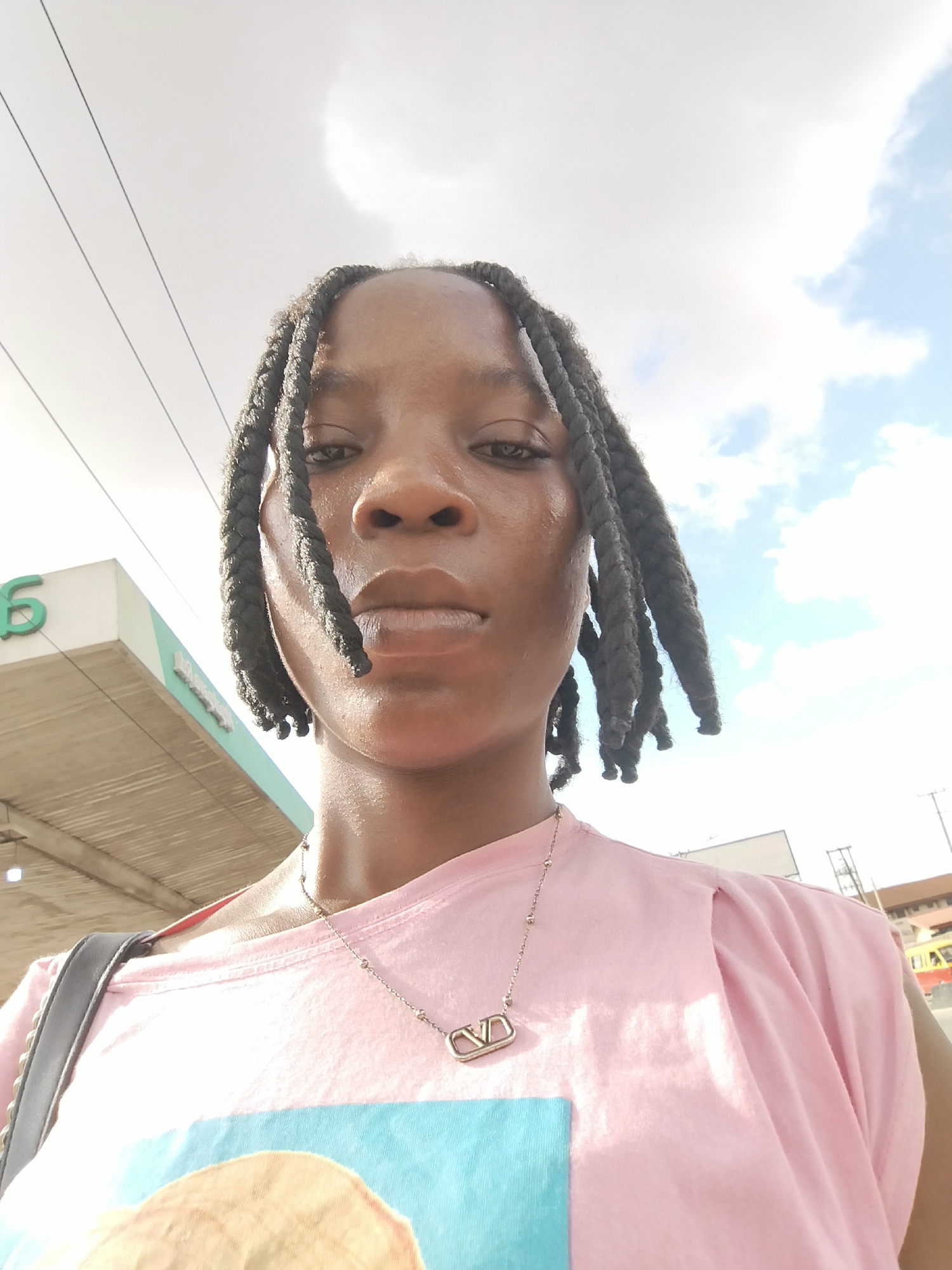 A young woman with braided hair takes a selfie outdoors. She is wearing a pink t-shirt and a silver necklace with a pendant. Behind her, a gas station canopy with a green "S" logo is visible, along with a cloudy sky. The lighting suggests it is daytime. The overall mood is neutral.