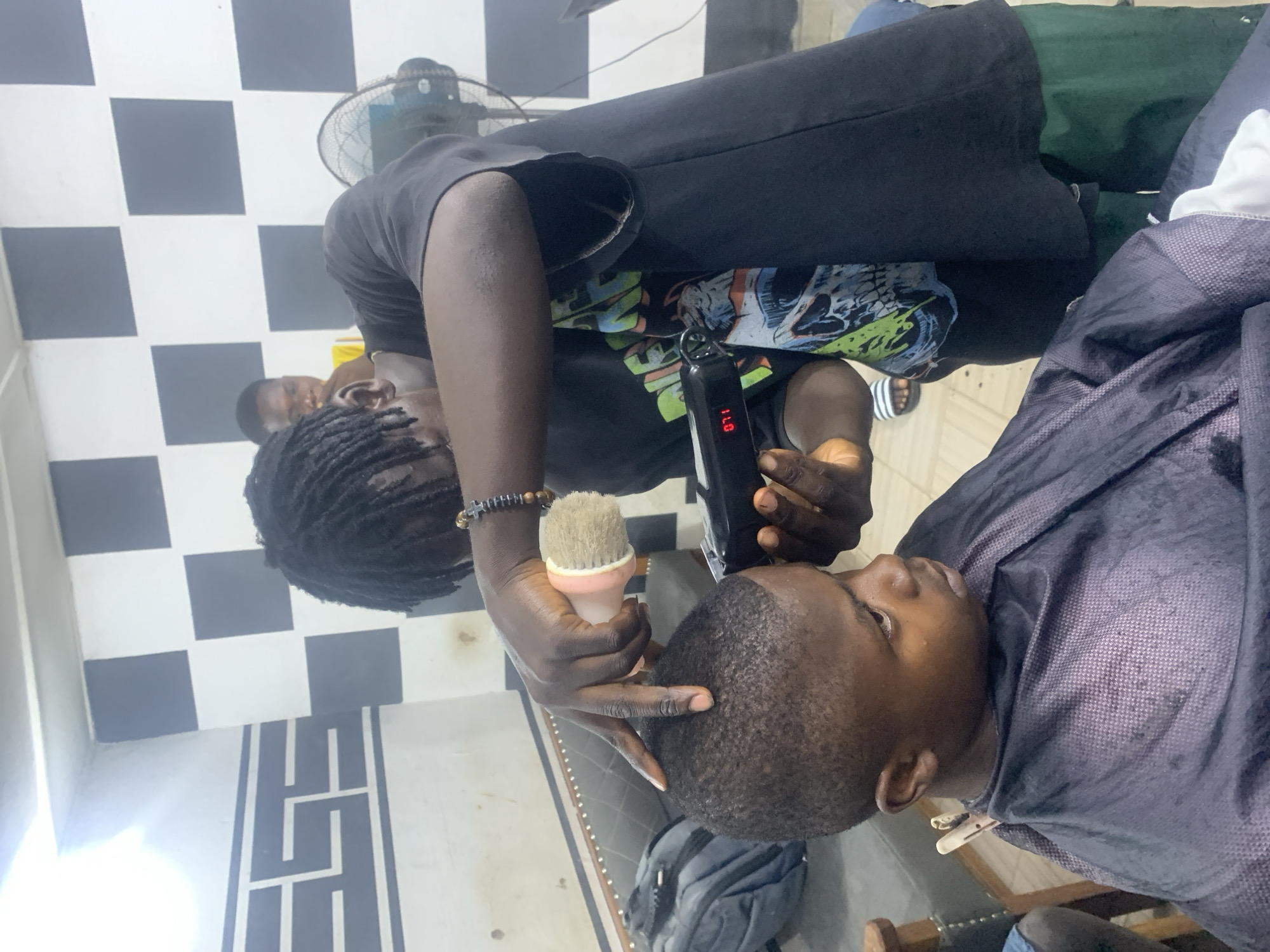 A barber with dreadlocks, wearing a black t-shirt and a beaded bracelet, is cutting the hair of a seated man wearing a blue barber cape. The barber is holding electric clippers in his right hand and a small brush in his left. The backdrop includes a black and white checkered tile floor and a black and white patterned wall near the floor. There is also another man in the background. The location is a barber shop in Jos, Nigeria.