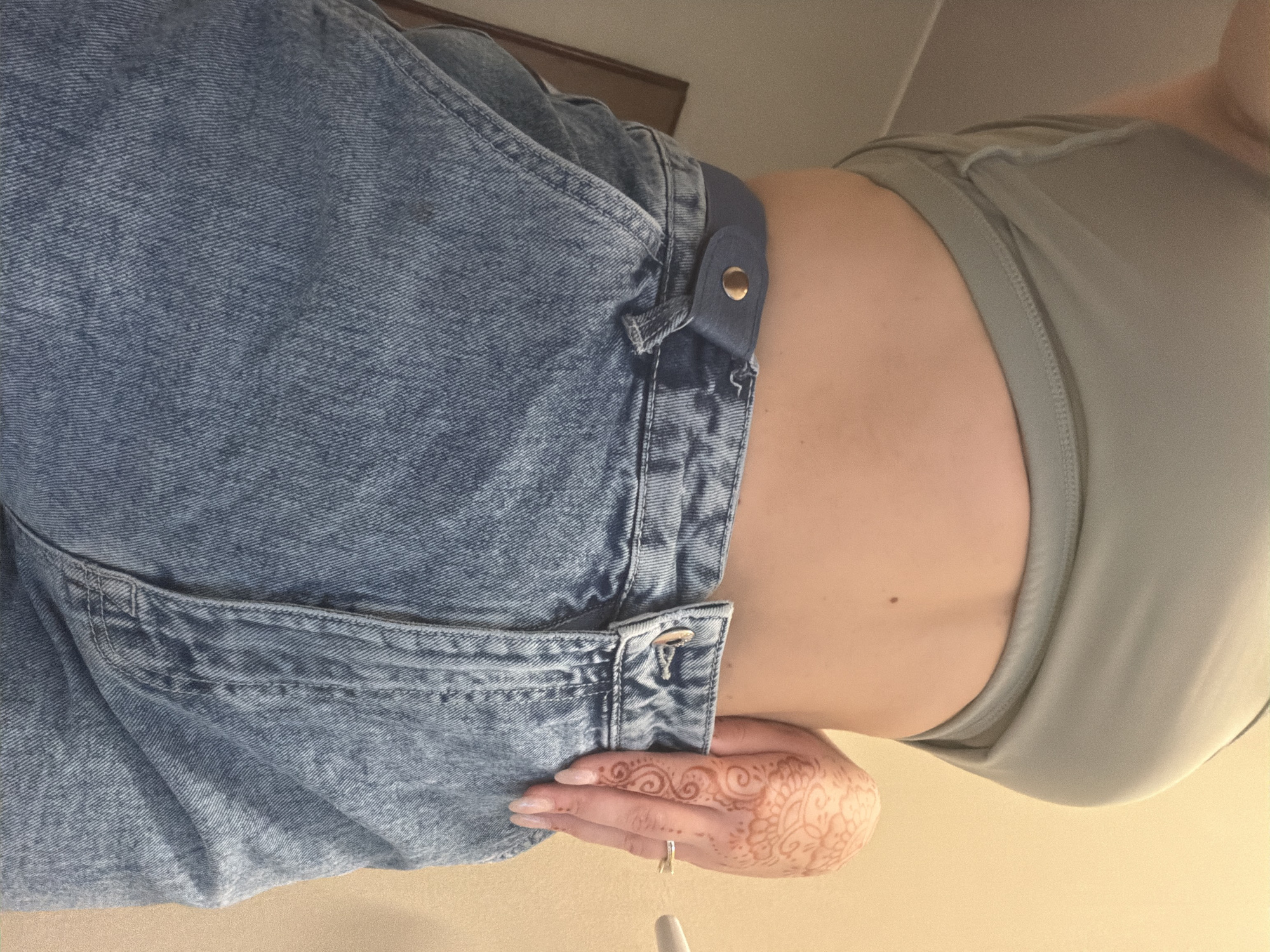 The image shows a close-up, low-angle view of a person wearing a light green crop top and high-waisted denim jeans. The person's midriff is exposed. They have a henna tattoo on the back of their right hand and are wearing a ring on their ring finger. The background is a plain, light-colored wall. The scene appears to be indoors, likely in a residential setting. There is no visible indication of the location being in Sofia, Bulgaria.