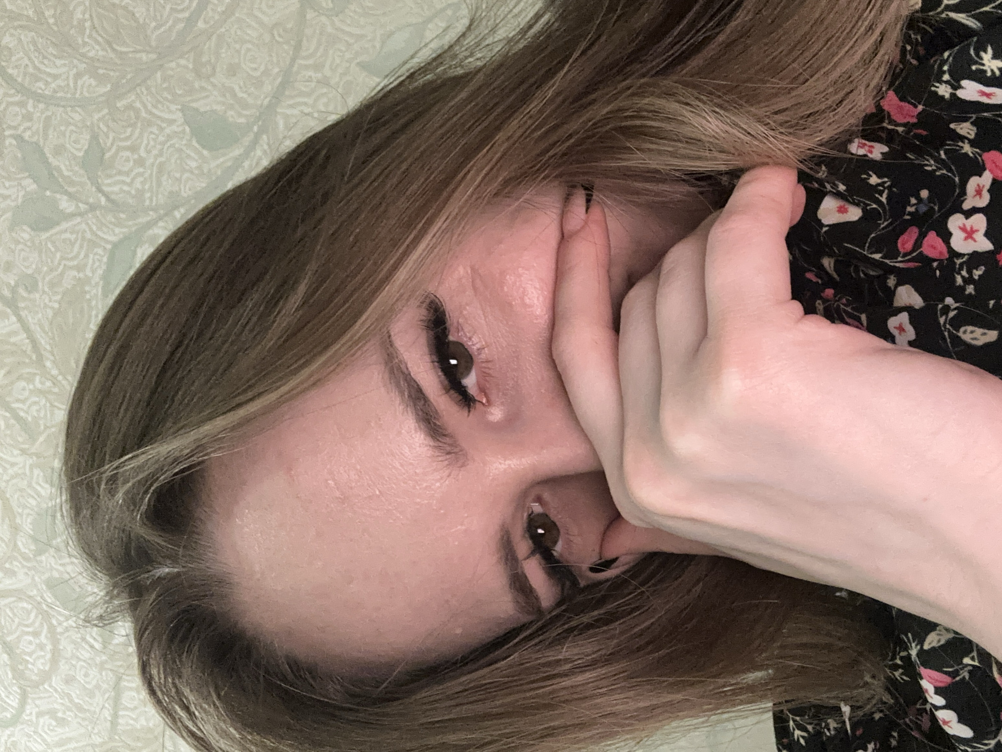 A close-up, selfie-style shot shows a young woman's face from the nose up. She has long, light brown hair that frames her face and falls over her right shoulder. Her left hand is brought up to cover her mouth, with her thumb tucked under her chin and her fingers obscuring her lips and part of her nose. She is wearing false eyelashes and has brown eyes that are visible peeking from behind her hand. Her skin appears flushed and slightly oily. The background is a textured, light-colored wallpaper with a faint, repeating pattern. The woman is wearing a black garment with a floral pattern of small pink and white flowers. The lighting is soft and diffuse.