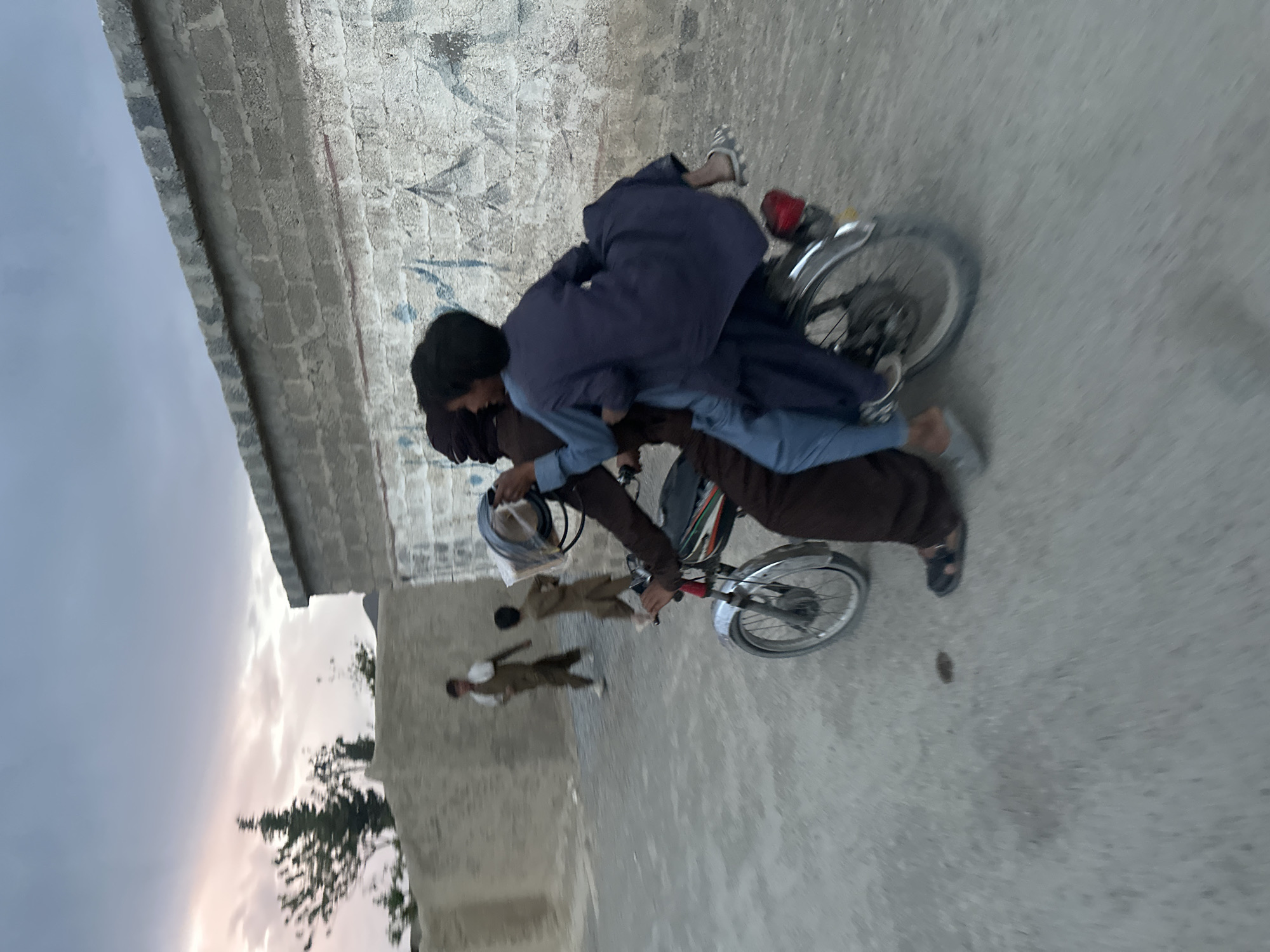 A man in brown clothing and a motorcycle helmet is riding a motorcycle with a young person lying across his lap in an outdoor urban environment. Two young children are standing nearby. The setting includes a brick wall and a partly cloudy sky.