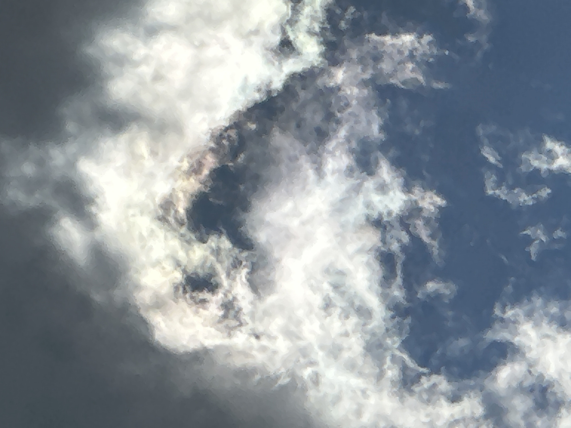 The image displays a detailed view of the sky in the city of Unknown, Unknown, characterized by a dynamic arrangement of clouds against a blue background. The lower and lower-right portions of the frame reveal a clear, deep blue sky. The central and upper areas are predominantly occupied by cloud formations. These clouds exhibit significant variation in illumination, with expansive bright white masses indicative of direct sunlight. Interspersed with and adjacent to these luminous clouds are darker, grey-toned cloud segments, most notably in the upper-central region, which create sharp visual contrast. Subtle iridescence, appearing as faint pink and light yellowish hues, is discernible along the edges of some brightly lit clouds, particularly where they abut darker sections. The cloud structures possess a textured, billowy morphology. No human subjects, identifiable man-made objects, or specific actions are present within the captured scene.